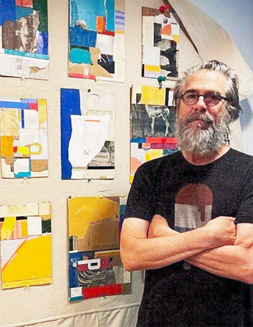 Man with gray hair and beard wearing glasses and a black T-shirt standing with arms crossed in front of colorful abstract artwork on the wall.