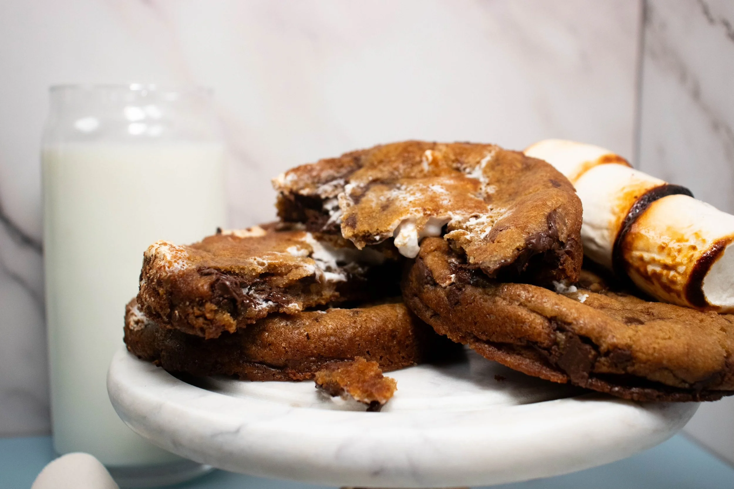 Close-up of a white marble cake stand with chocolate chip cookies and a chocolate-dipped wafer roll, with a glass of milk in the background.