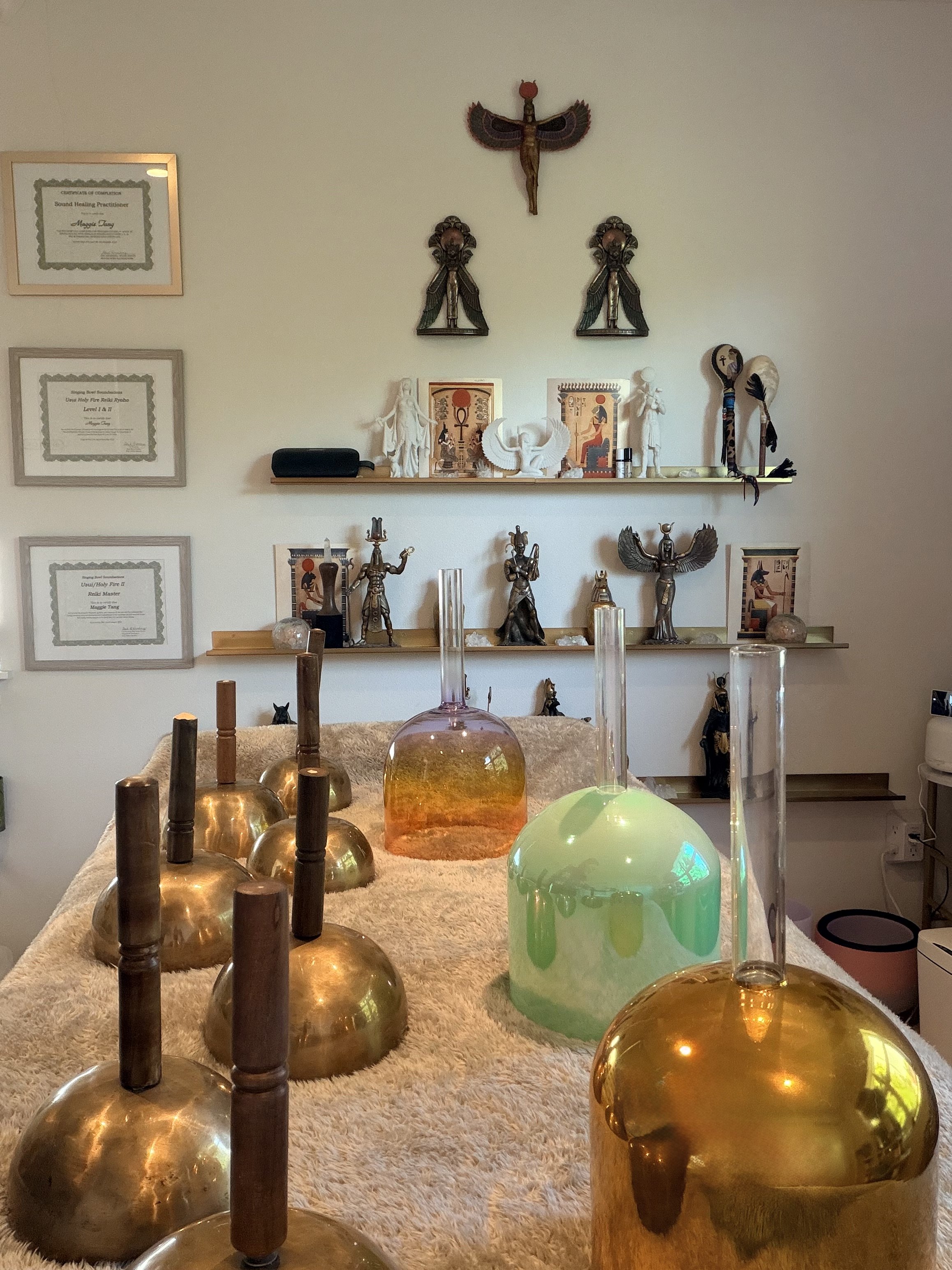 Interior view of a room with spiritual and spiritual artifacts. Decor includes framed certificates on the wall, Egyptian-style statues, and Alex Grey artwork. On a table in the foreground, there are multiple brass bells and colorful glass objects, including a large orange glass vessel and a green glass sculpture. Behind, shelves hold various statues, artifacts, and artwork, including depictions of Egyptian gods and other figures.