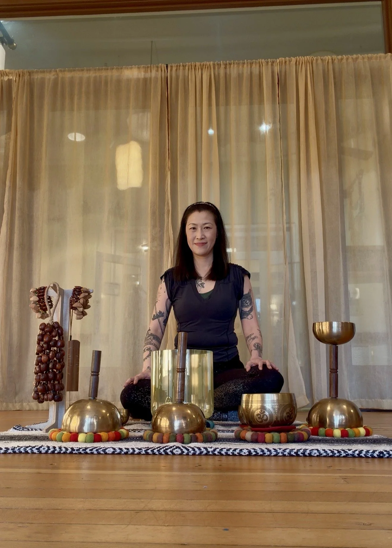 Woman sitting cross-legged on a patterned rug surrounded by musical instruments, including singing bowls and decorative items, in front of a sheer beige curtain.