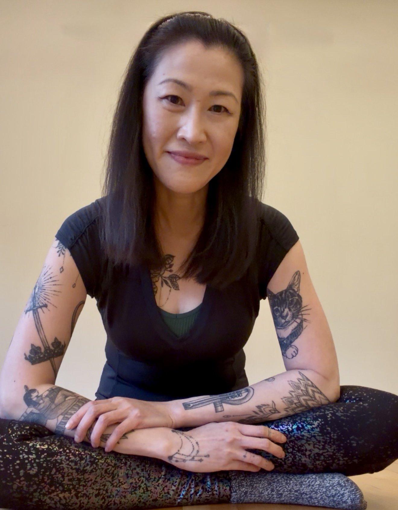 A woman with shoulder-length dark hair and tattoos on her arms sitting cross-legged on a patterned cushion, smiling at the camera.