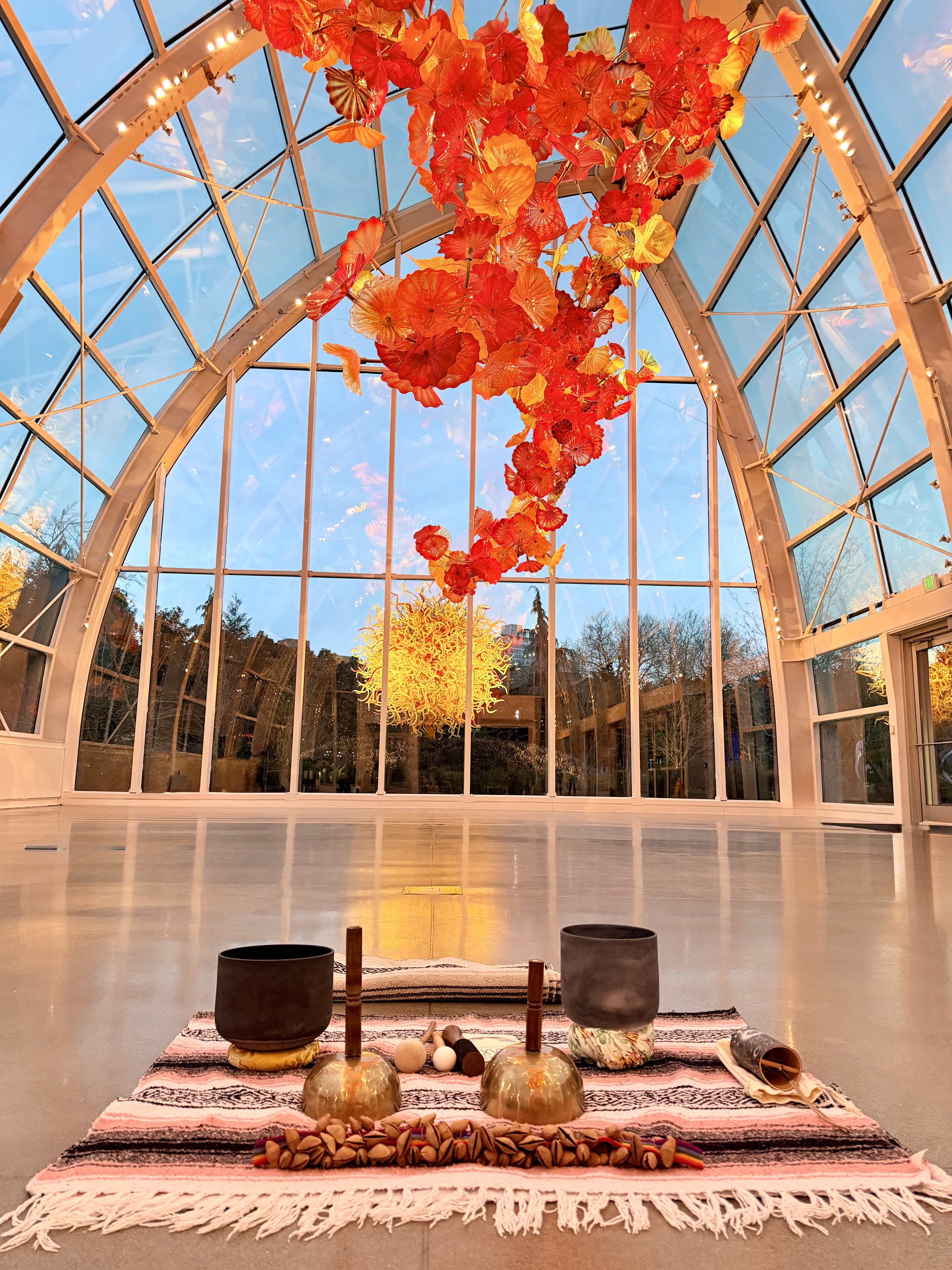 Interior view of a greenhouse with a high glass ceiling and walls, decorated with a hanging art installation of orange and yellow glass sculptures resembling leaves or flowers. On the floor, there is a striped rug with objects including singing bowls, wooden sticks, rocks, a rolled-up blanket, and other spiritual or meditation items, suggesting a setting for meditation or ceremony.