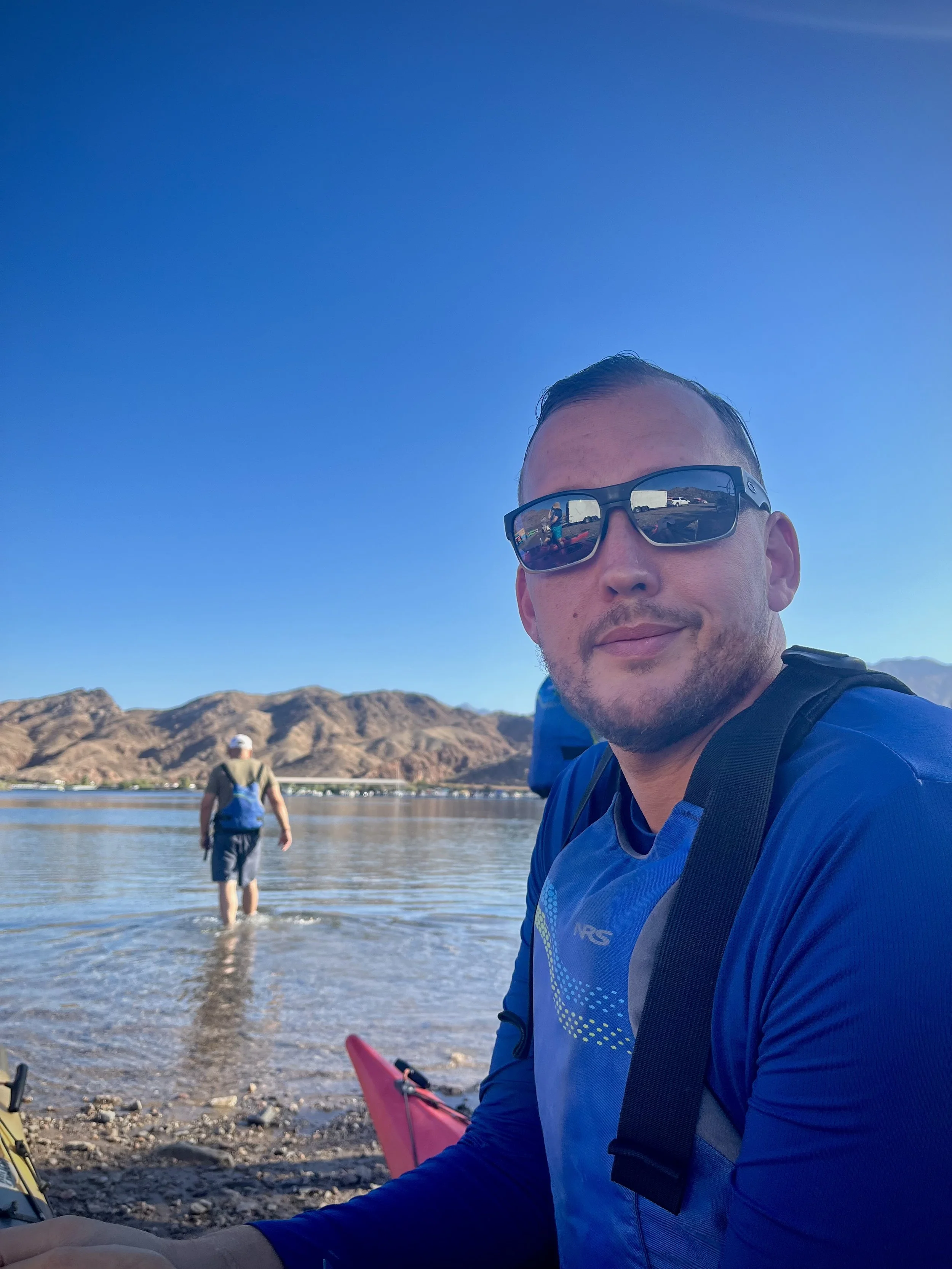 A man wearing sunglasses and a blue jacket taking a selfie near a body of water with mountains in the background. Another person is walking in the water, and a pink kayak is partially visible.