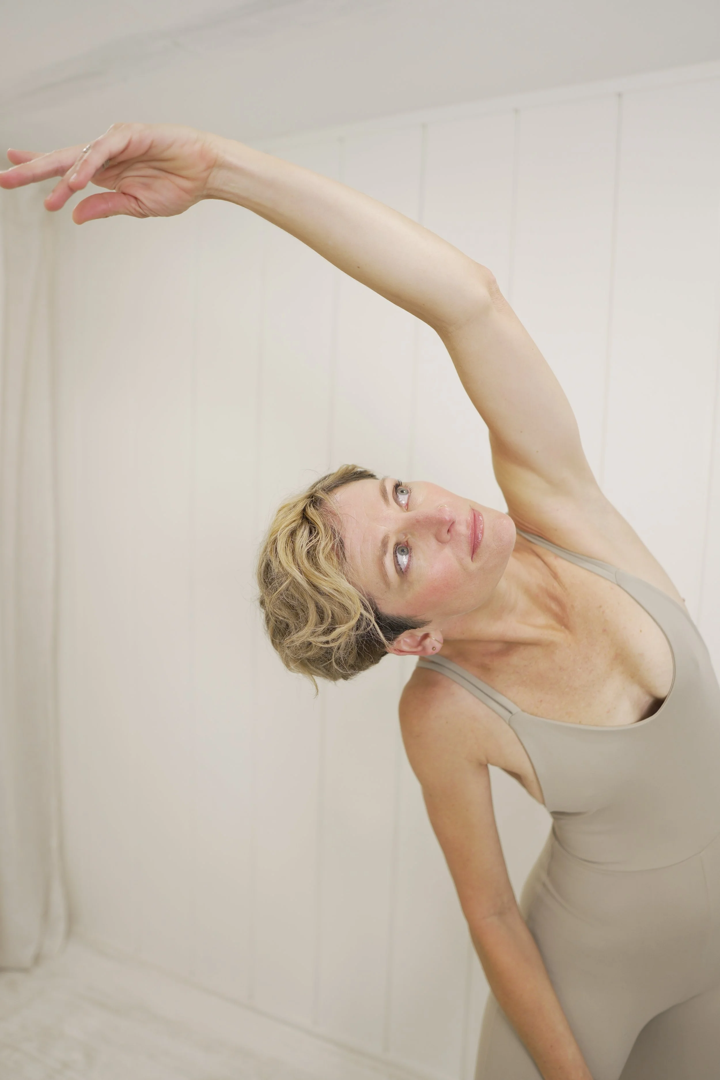A woman with blonde hair is practicing yoga or stretching in a room with white paneled walls. She is wearing a beige tank top and beige yoga pants, with her right arm extended upward and her gaze directed toward her hand.