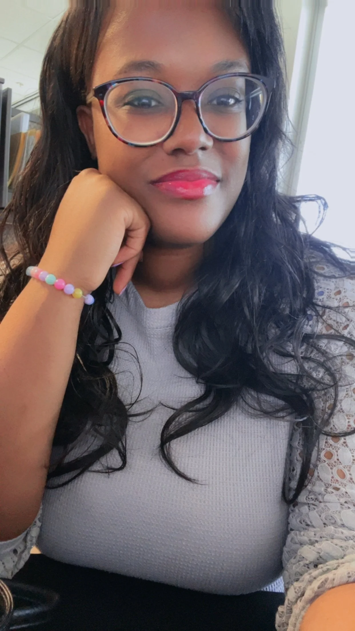 A woman wearing glasses with a black frame and a white shirt with lace on the sleeves, smiling softly with her hand resting on her chin, and a colorful beaded bracelet on her wrist.