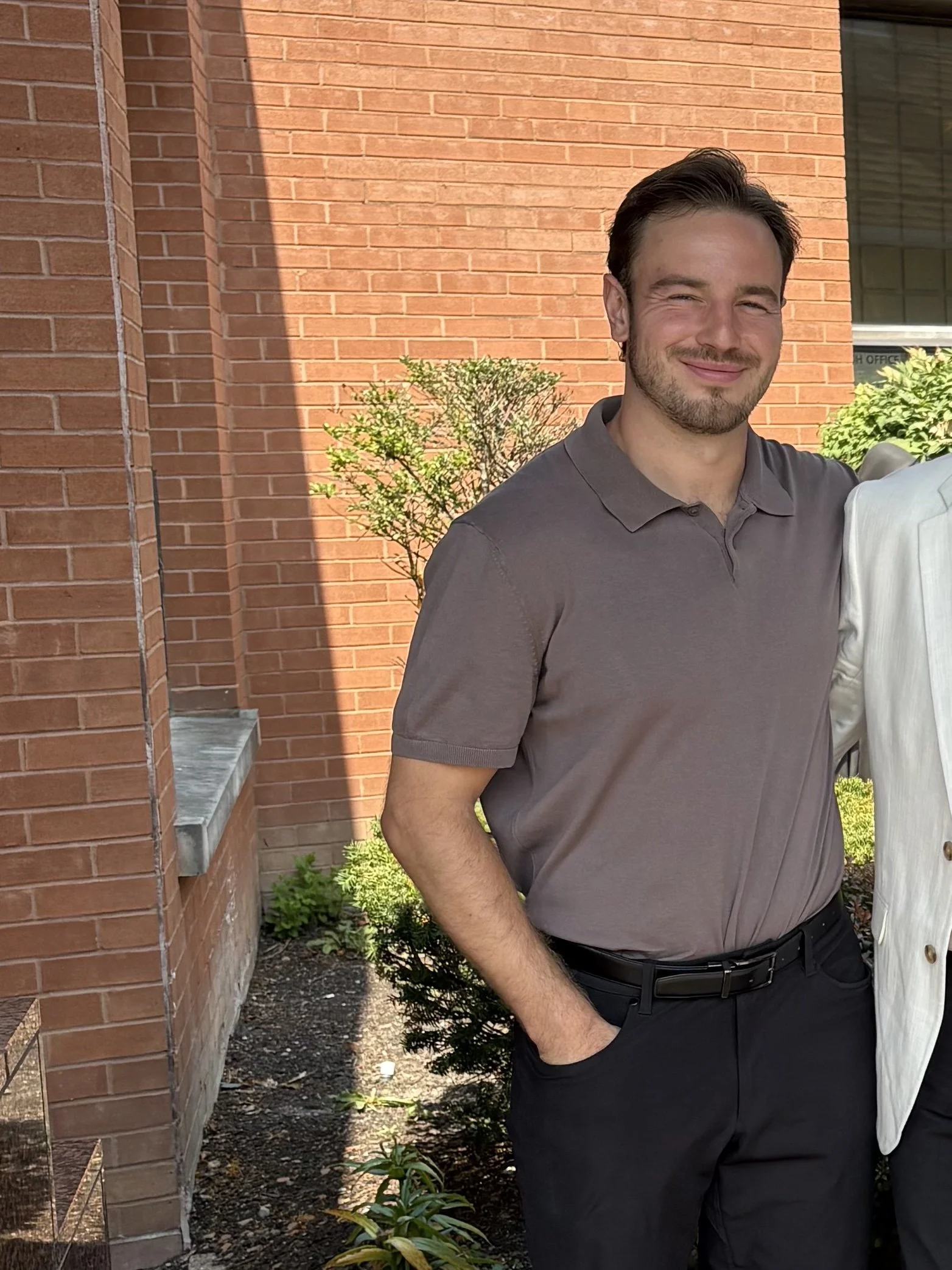 A man with dark hair, a beard, and light skin, smiling and standing outside near a brick building, wearing a gray polo shirt and black pants.
