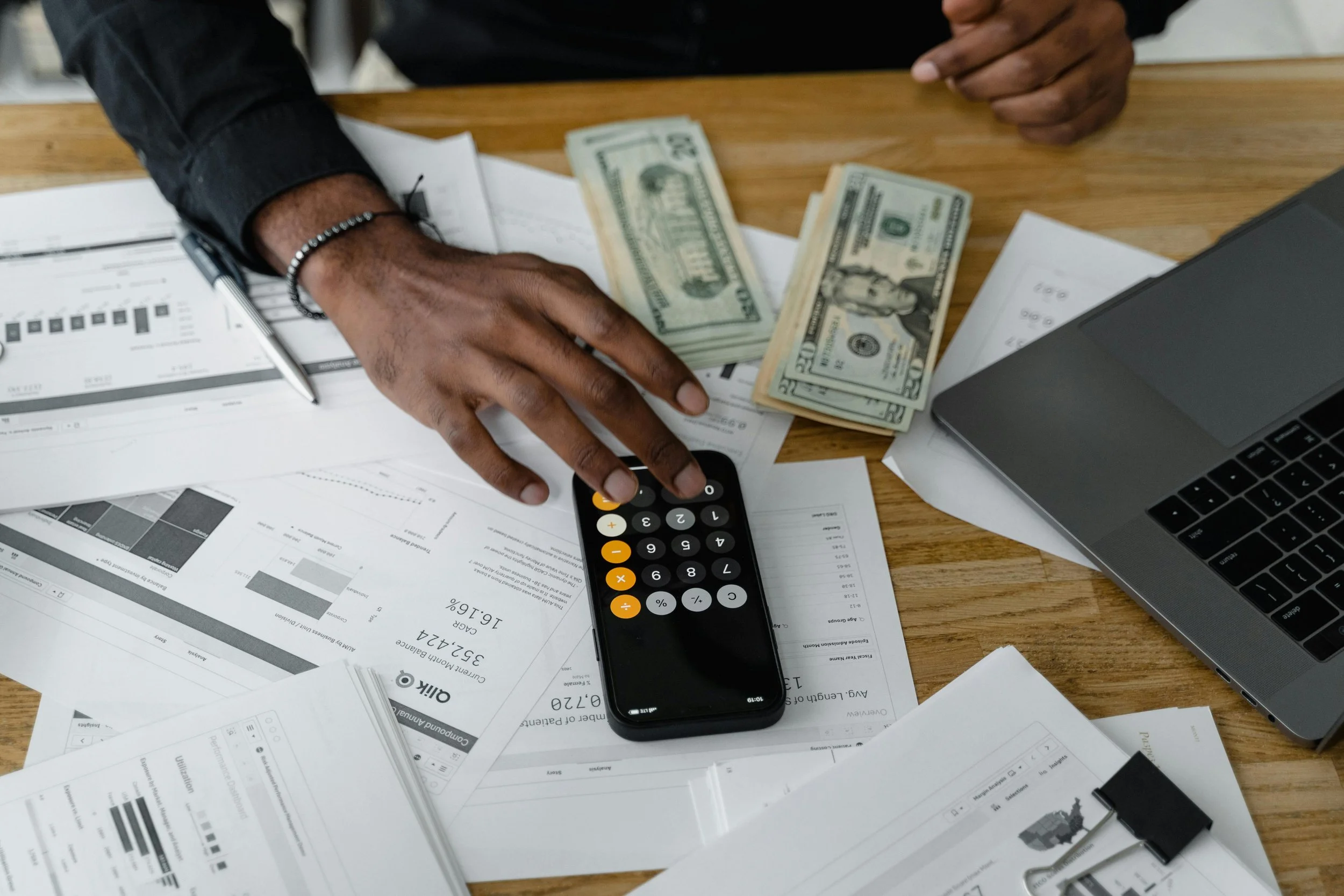 A person handling cash, stacks of twenty-dollar bills, a calculator, a pen, a laptop, and scattered papers with graphs and documents on a wooden desk.