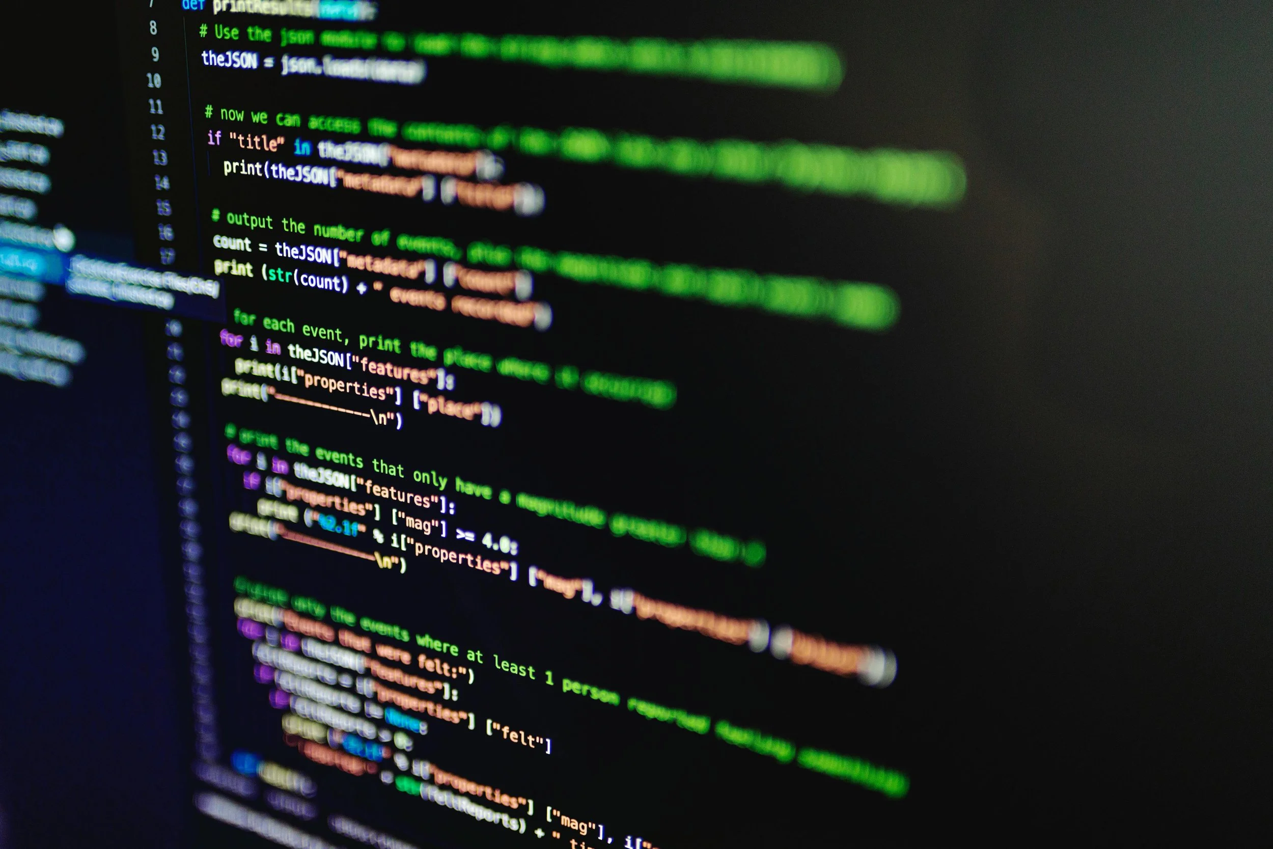 Close-up of a computer screen displaying lines of colorful programming code in various colors like green, purple, blue, and orange on a dark background, with some text blurred.
