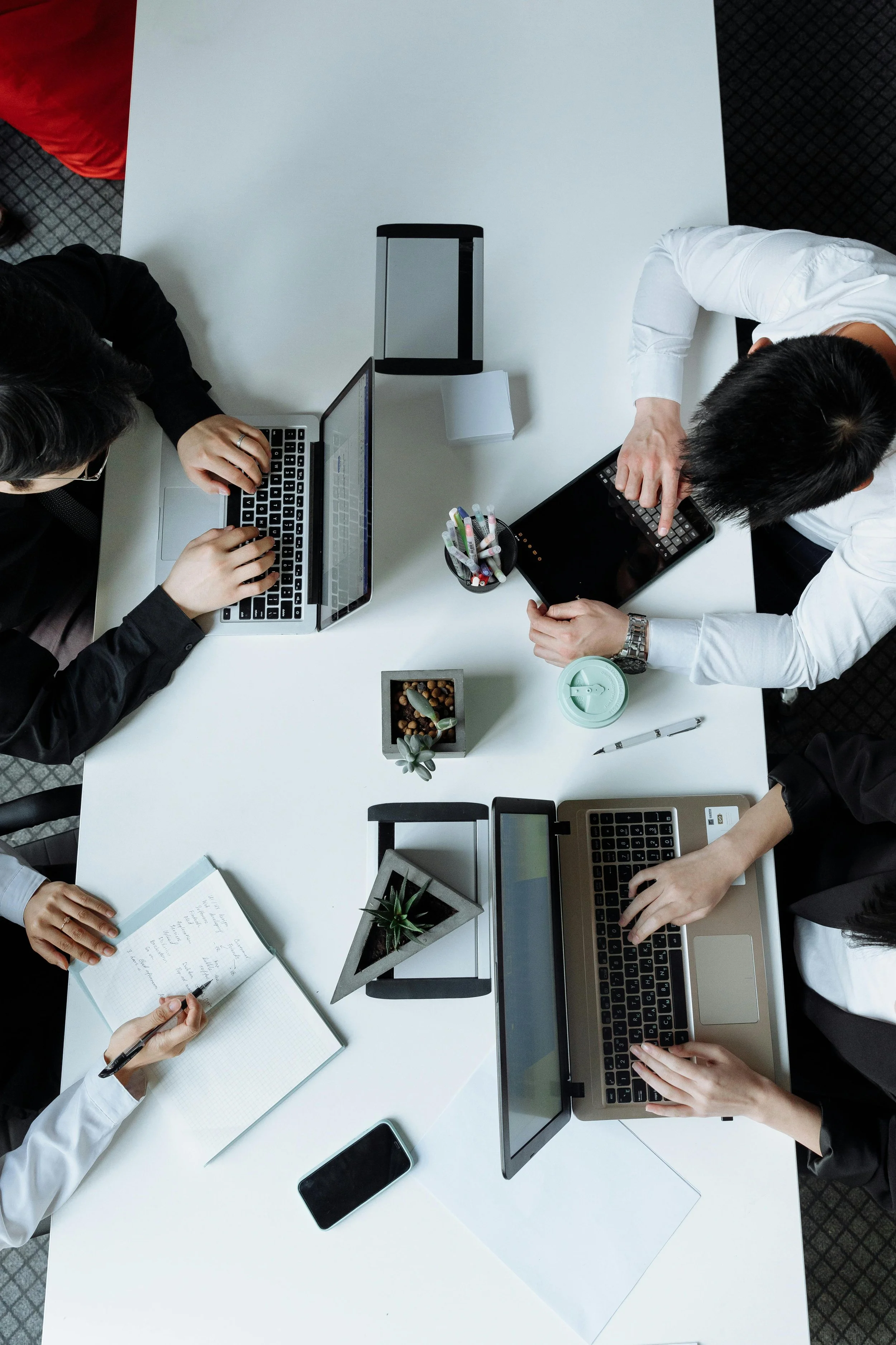 Top-down view of a group of people working at a white conference table with laptops, a tablet, notebooks, pens, a small plant, a container of markers, and a cup with a lid.