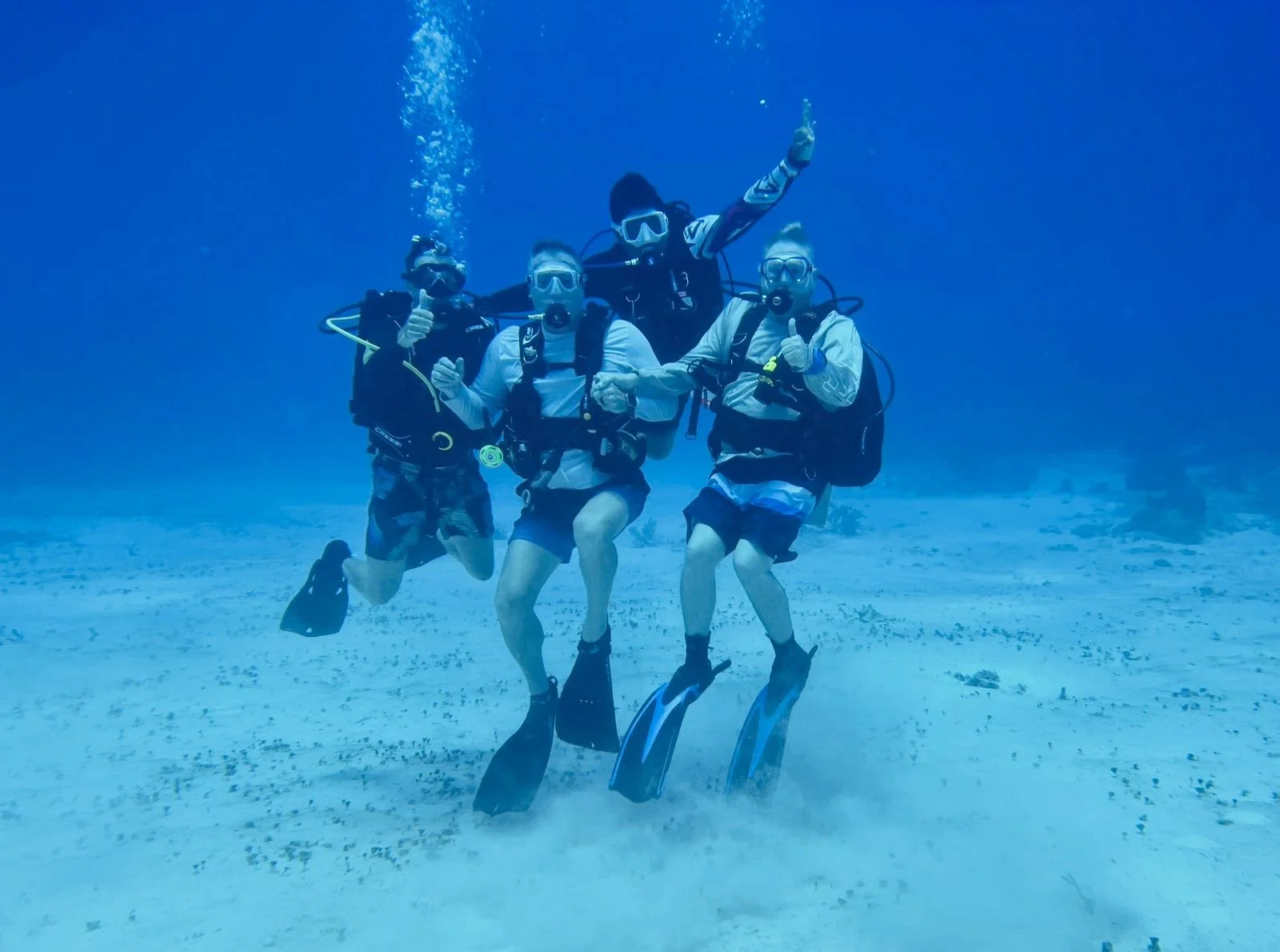 Four people in scuba gear underwater, posing for a photo, giving thumbs up, and one making a peace sign.