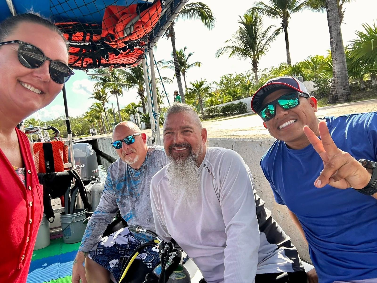 Four people smiling for a selfie outdoors in a tropical setting with palm trees. They are dressed casually, and one is making a peace sign. Two men are seated, one woman and another man are standing.