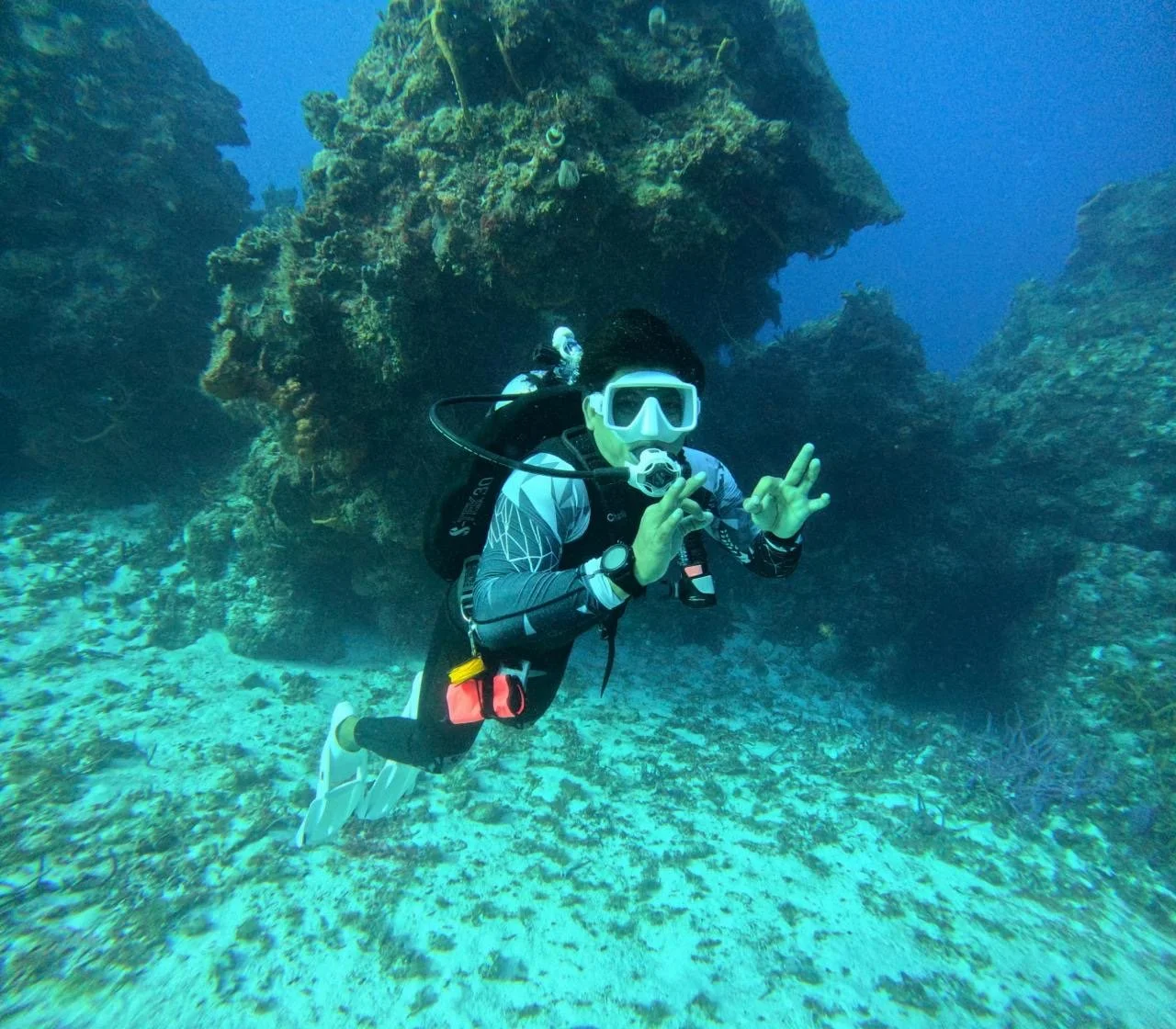 A scuba diver underwater surrounded by coral formations making an 'OK' hand sign.