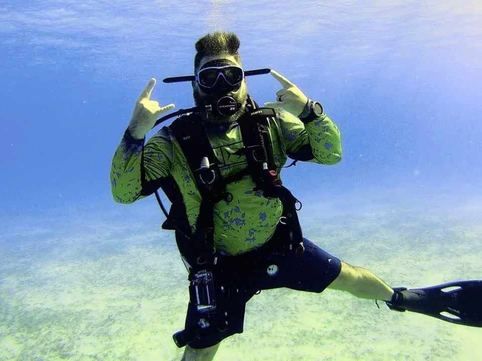 A man is scuba diving underwater, wearing a green shirt, black shorts, diving mask, and equipment, making a gesture with his hands.