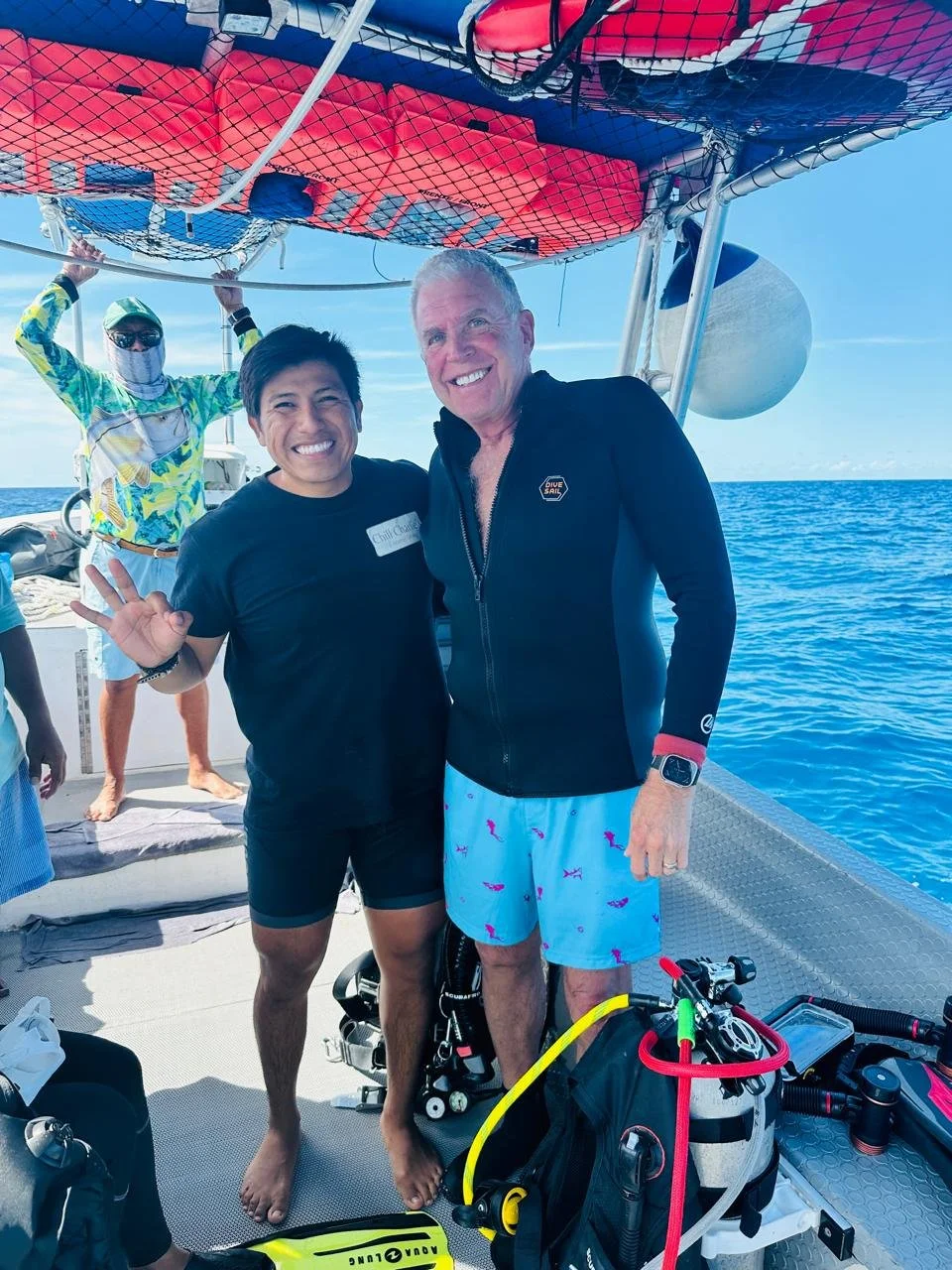 Two smiling men posing on a boat during a scuba diving trip, with scuba gear and the ocean in the background.