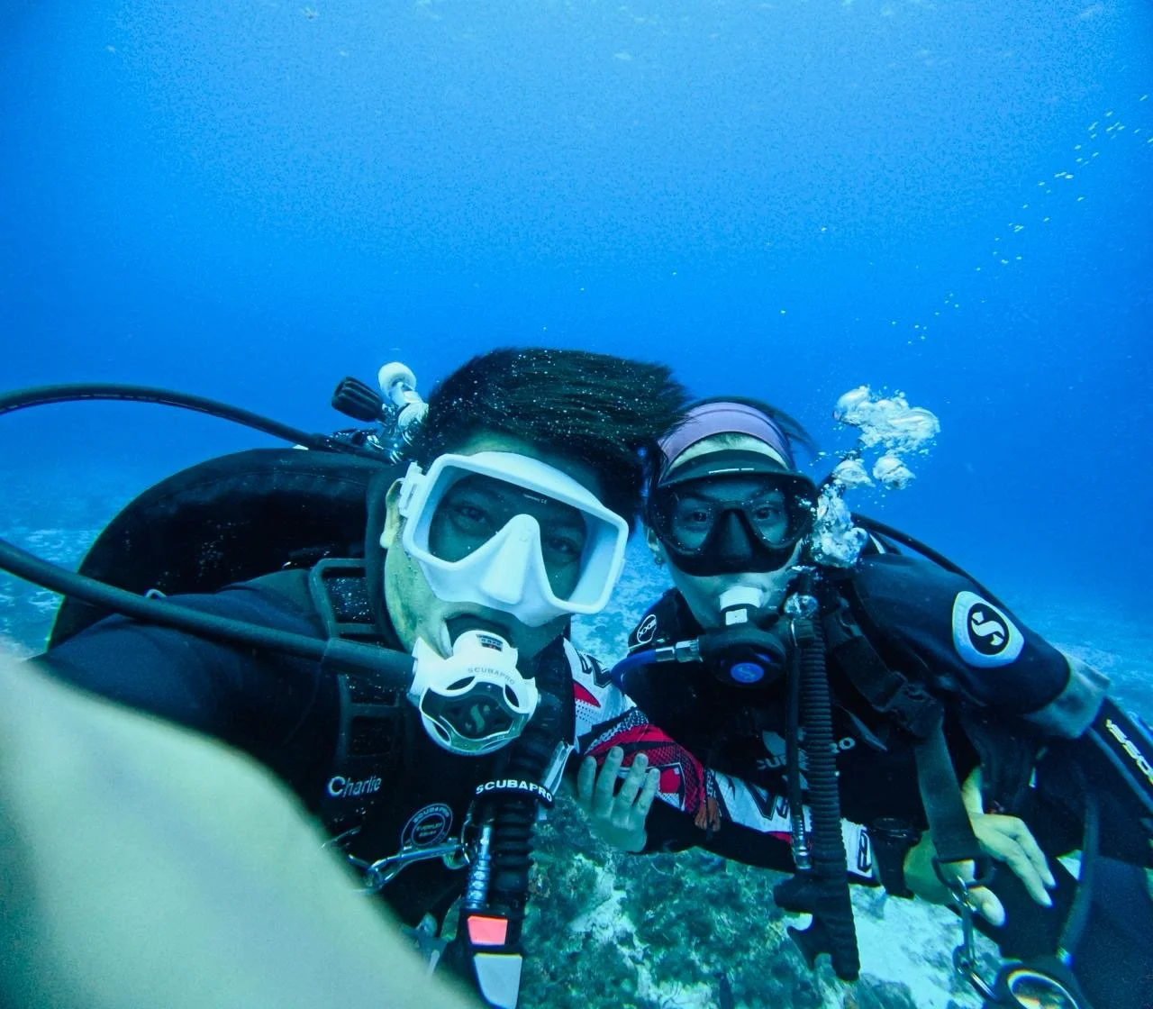 Two scuba divers underwater taking a selfie with coral and blue water in the background.