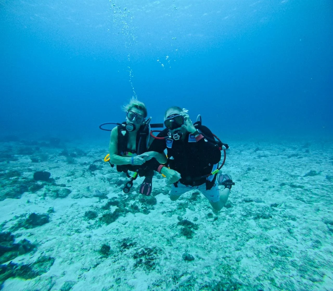 Two scuba divers underwater holding hands over a coral and rocky ocean floor.
