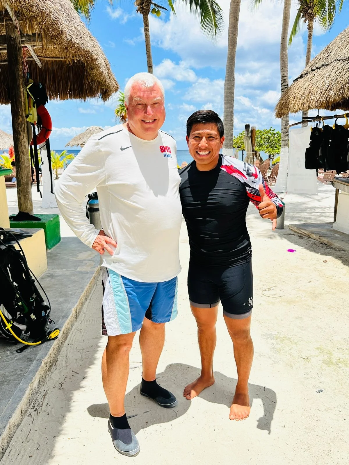 Two men posing on a beach under palm trees. One is wearing a white long-sleeve shirt and shorts, the other is in a black athletic shirt and shorts. They are smiling, with the man in black giving a thumbs up.