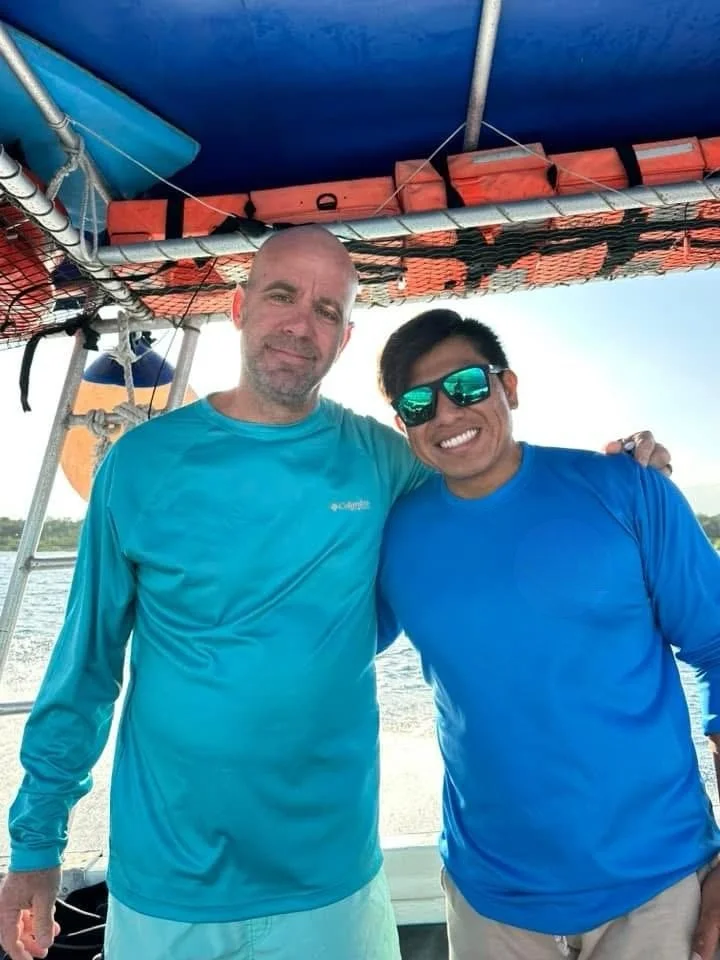 Two men wearing blue shirts smiling on a boat, with water and sky in the background.