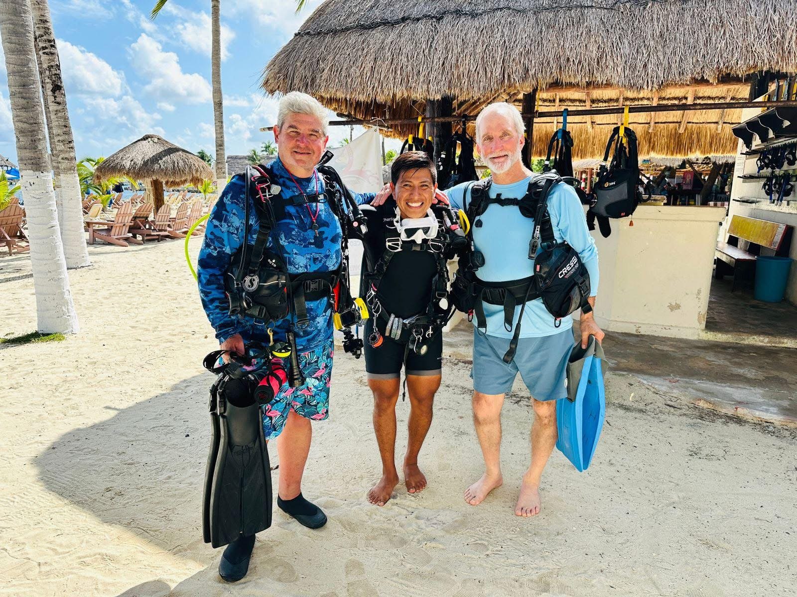Three people stand on a sandy beach with palm trees and beach chairs in the background, dressed in scuba diving gear, smiling at the camera.