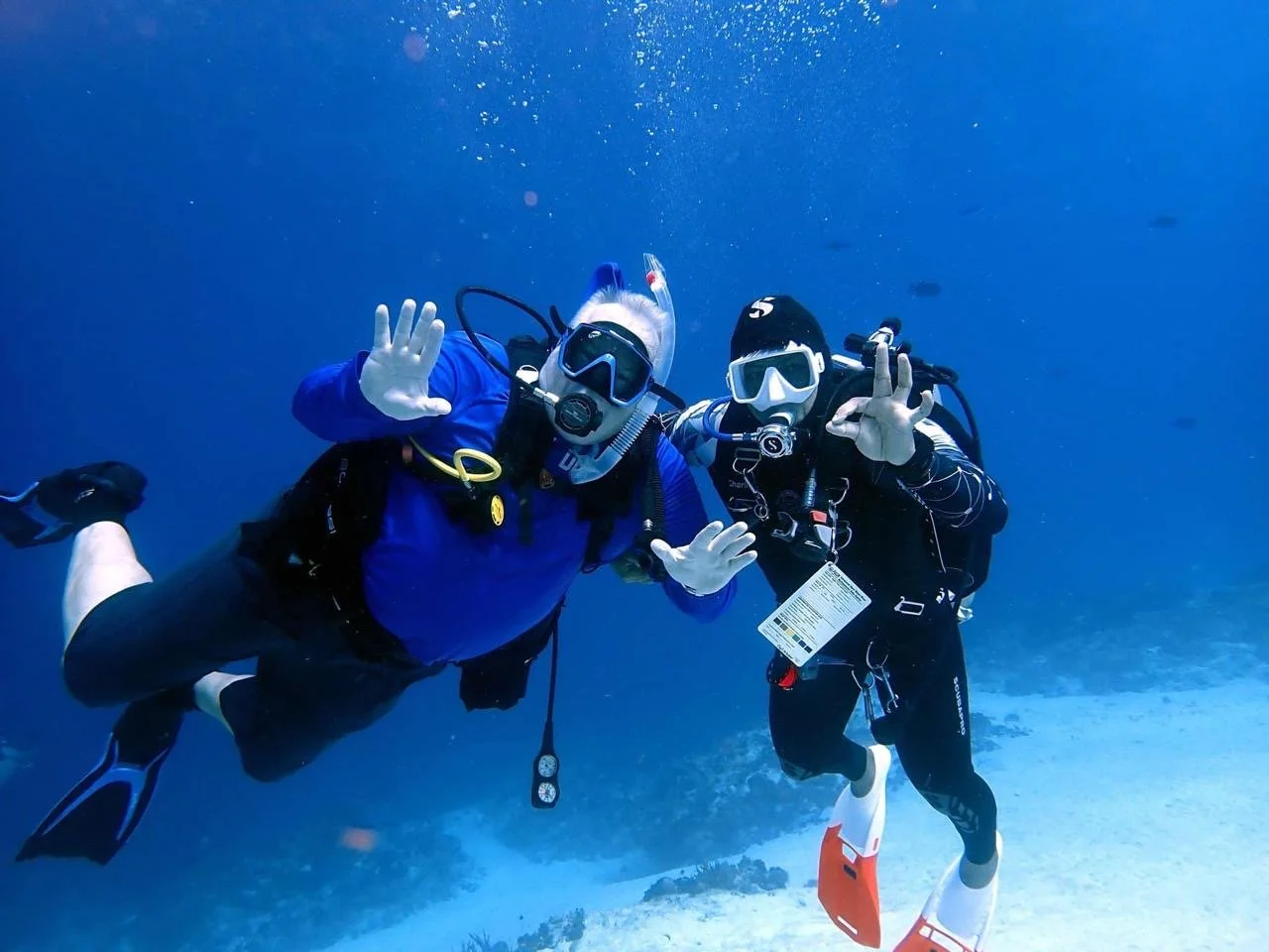 Two scuba divers in wetsuits and masks underwater, waving and making hand signs.