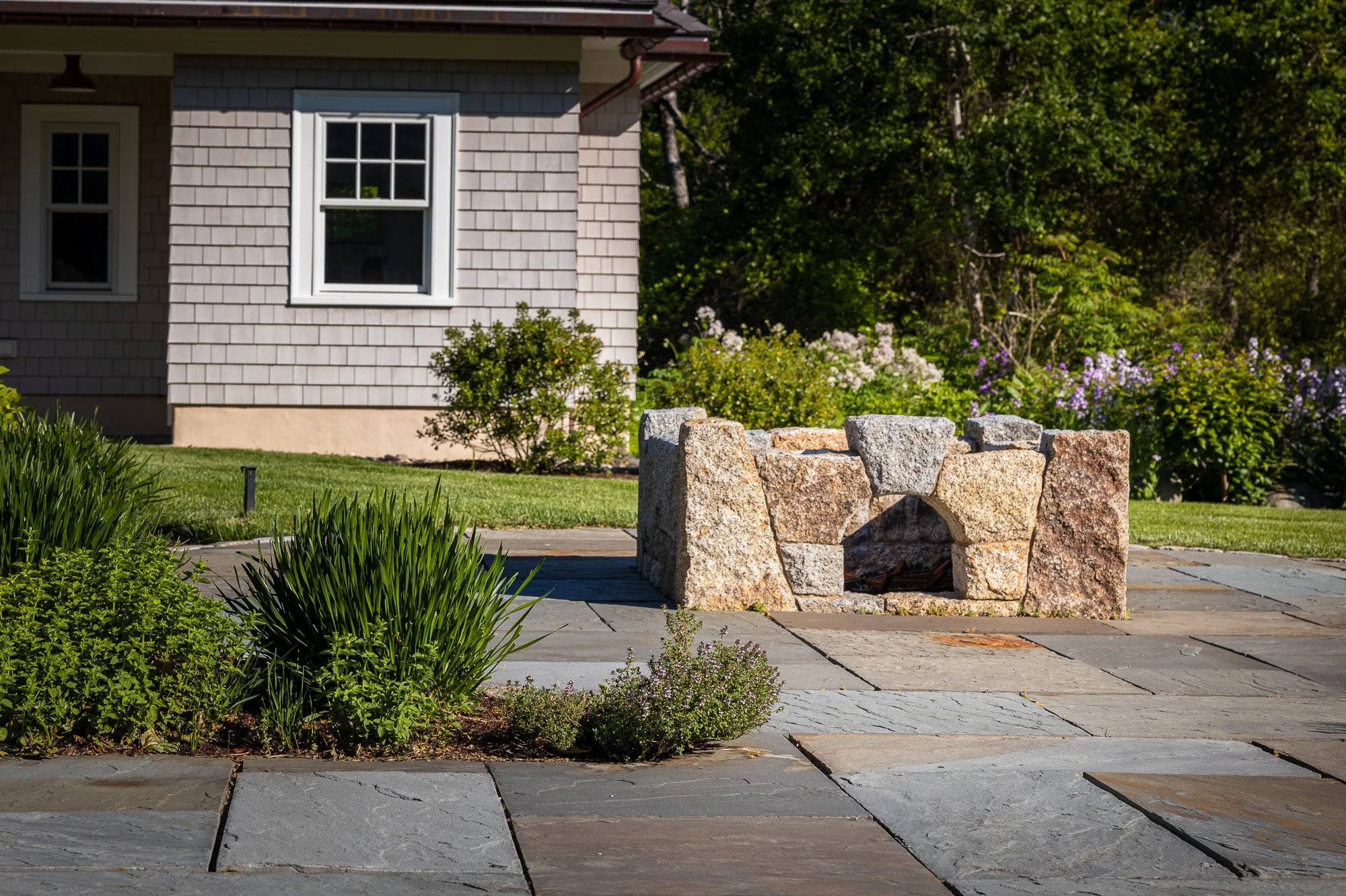 A house with grey wooden shingles, white window frames, and a garden with green bushes and purple flowers. In the foreground, there is a stone fire pit surrounded by a stone patio.