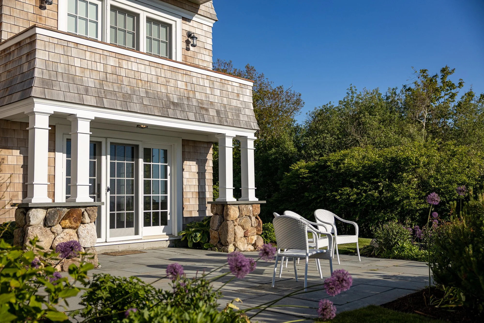 The image shows the exterior of a house with a small porch area, stone columns, and a patio with two white chairs. There are purple flowers in the foreground and green trees and bushes in the background under a clear blue sky.