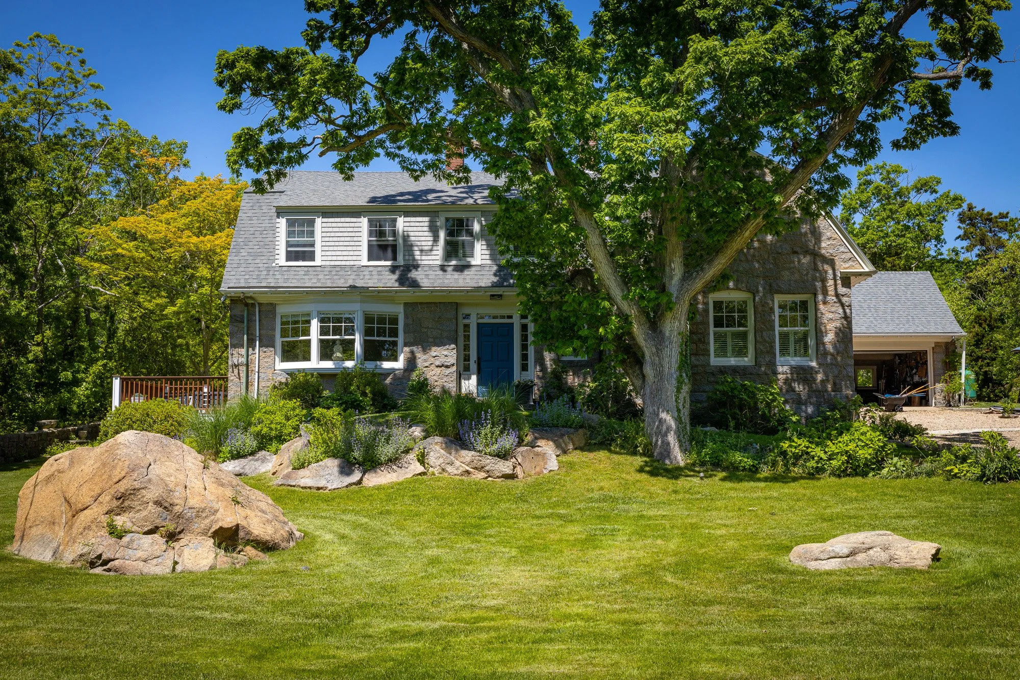 A gray stone house with a dark blue door, multiple white-framed windows, and a gray shingle roof surrounded by trees, rocks, and a well-kept lawn under a clear blue sky.