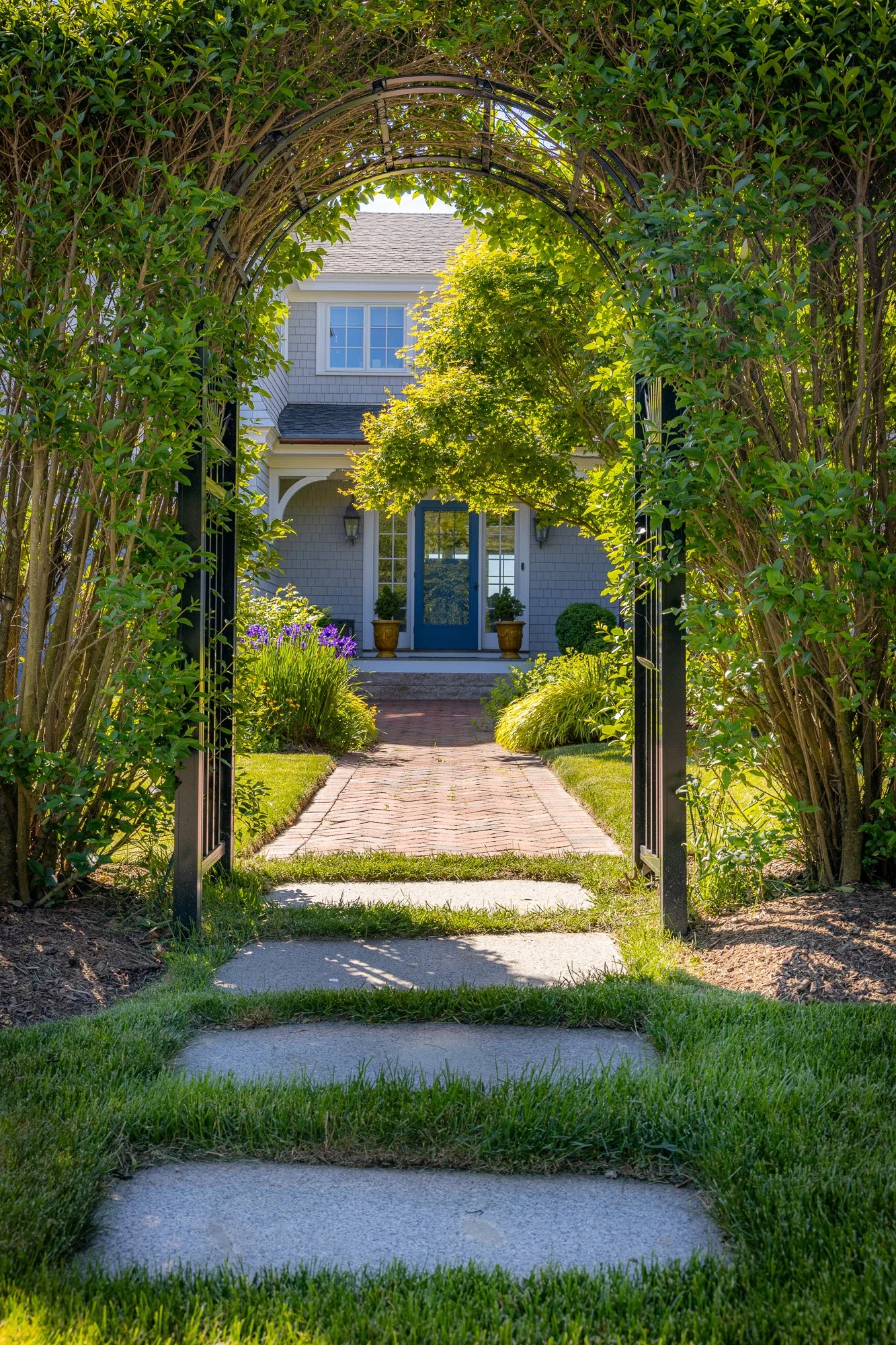View through a garden gate showing a brick pathway leading to a house with a blue door, surrounded by green trees, shrubs, and purple flowers.