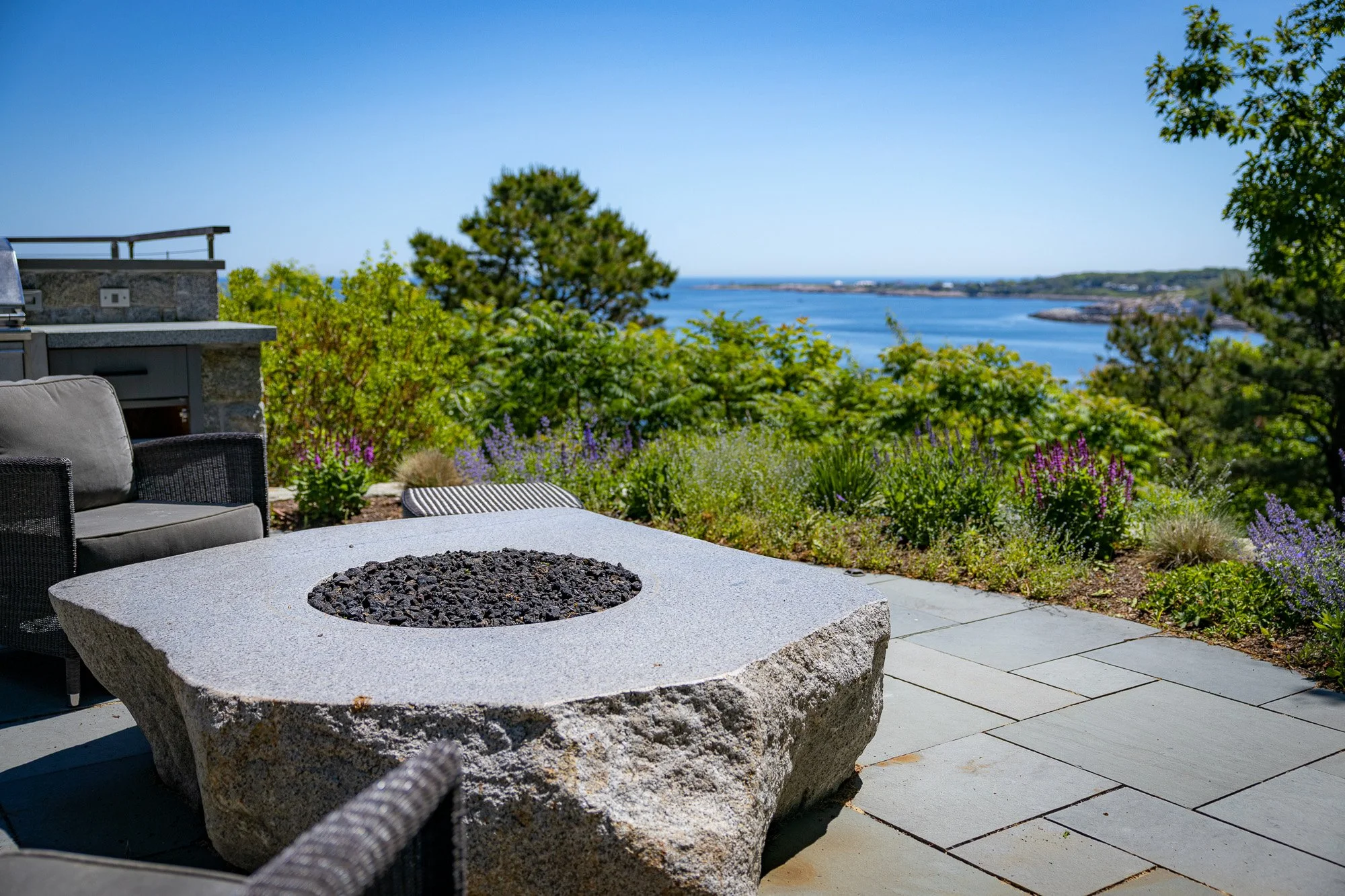 Outdoor seating area with a fire pit made of large granite, surrounded by a patio with potted plants and overlooking a scenic water view with trees and shoreline.