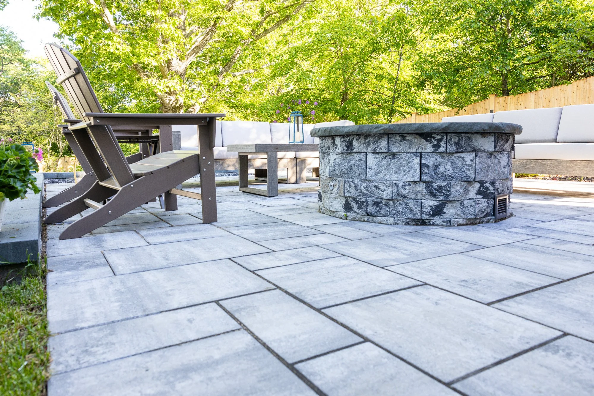 An outdoor patio with a stone fire pit, gray cushioned sectional sofa, and outdoor chairs surrounded by green trees and a wooden fence.