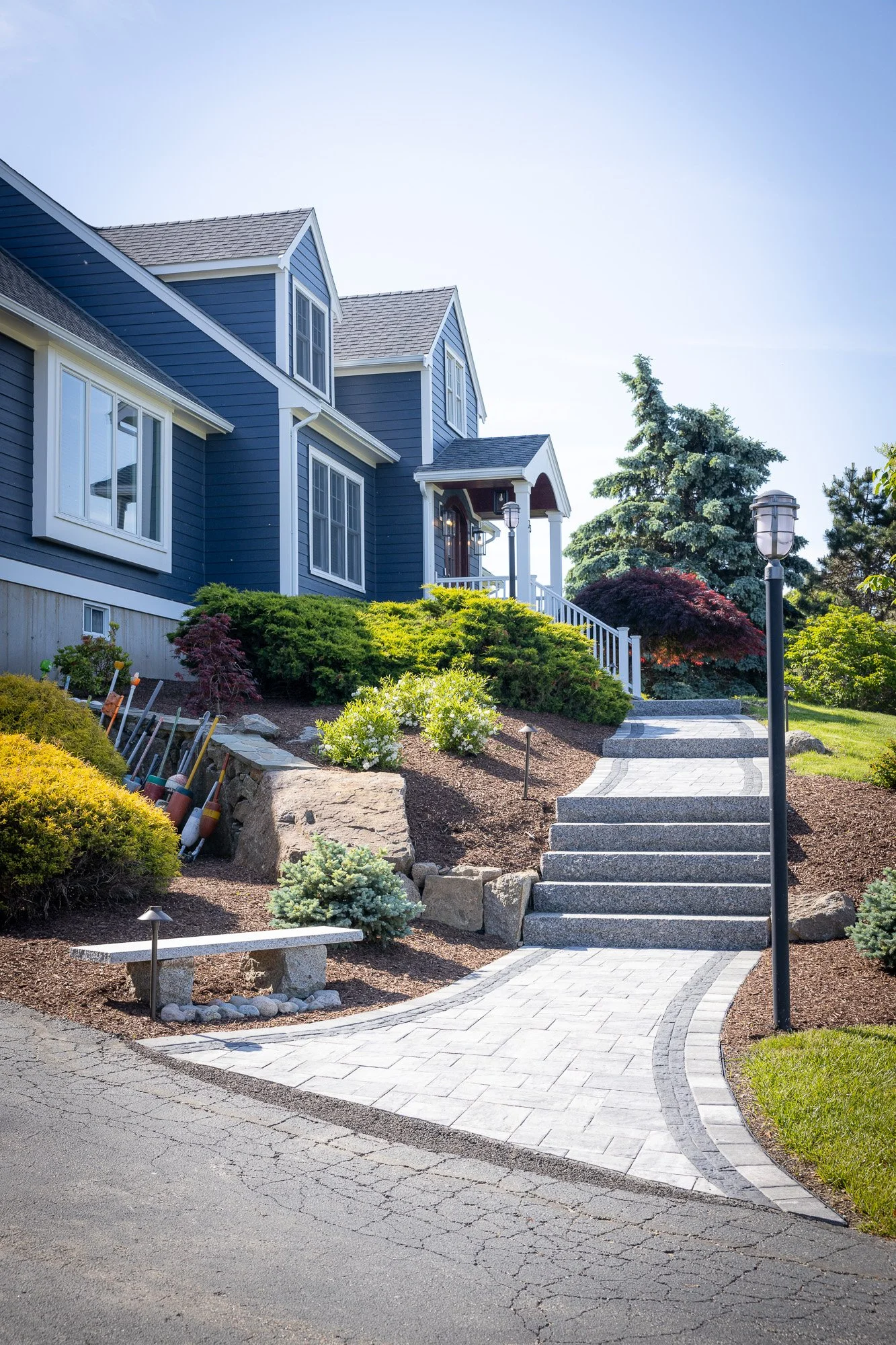 A landscaped front yard with a stone pathway leading to a house, garden beds, bushes, and trees, with exterior lighting fixtures.