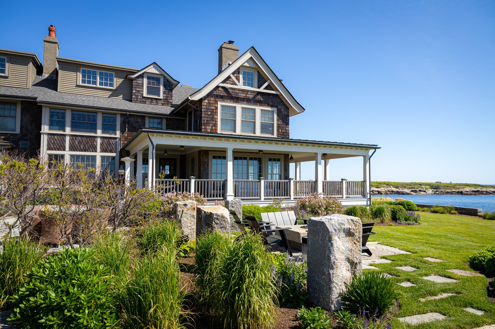A large house with brown shingles and white trim, featuring multiple gabled roofs and several windows overlooking a coastal landscape with a body of water. In the foreground, there is a garden with green plants, large rocks, a stone pathway, and outd