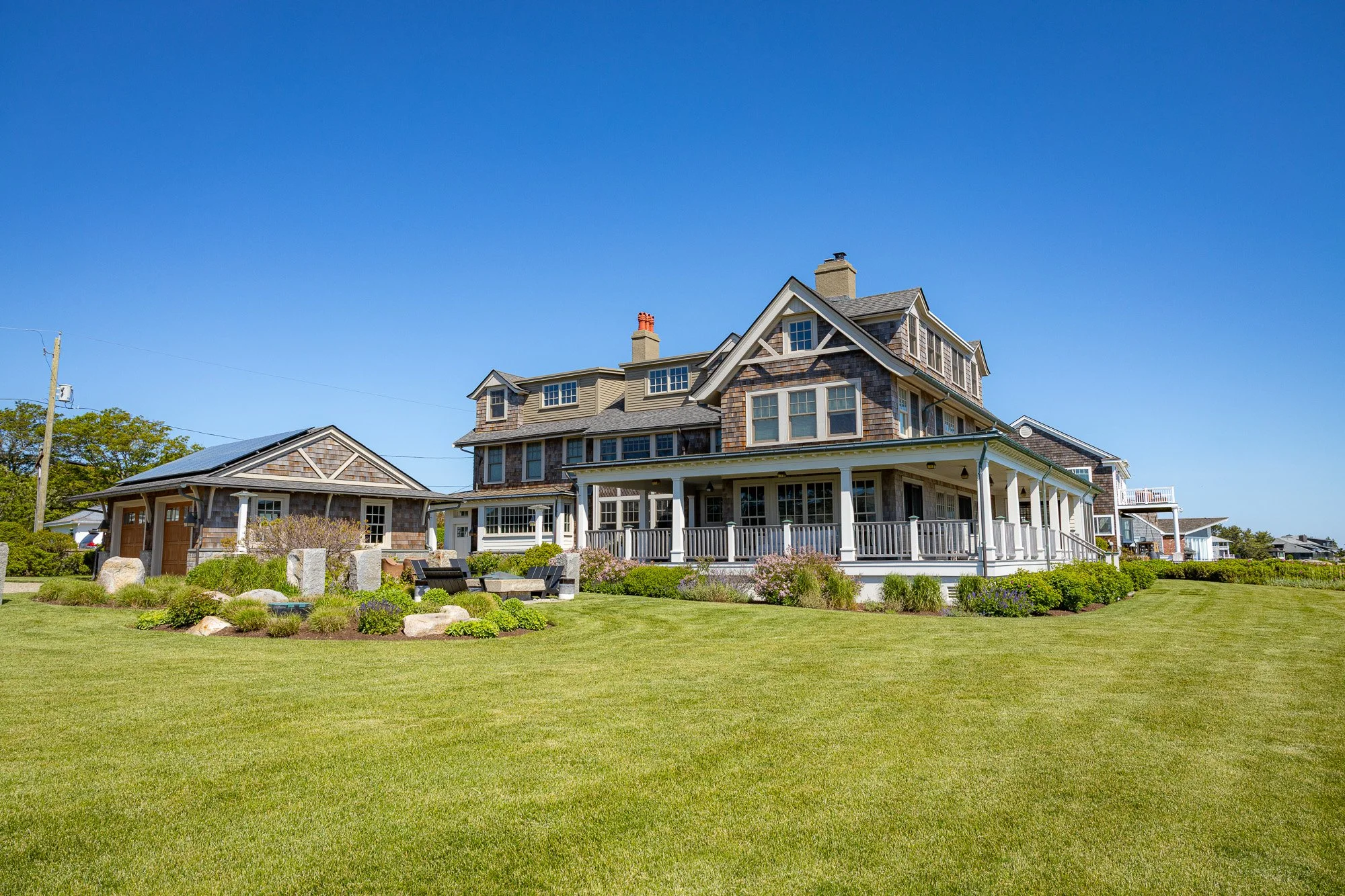 A large, multi-story house with a porch, surrounded by a well-maintained lawn and garden under a clear blue sky.