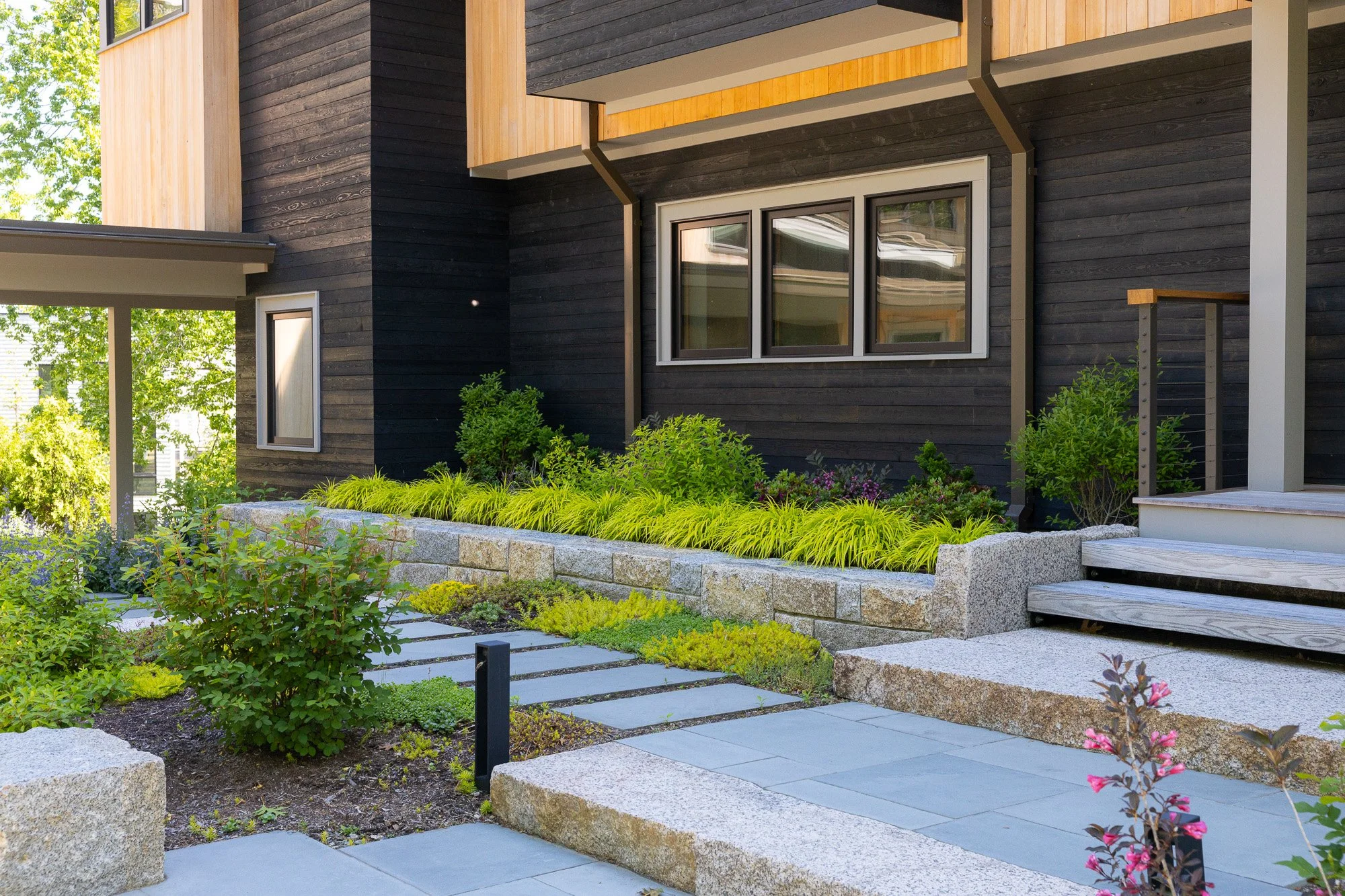 Modern house with black and wood exterior, surrounded by landscaped garden with bushes, flowers, and stone pathways.