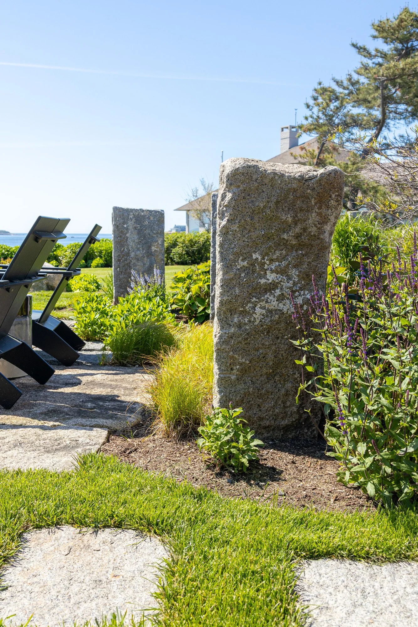 Outdoor garden with large rocks, green plants, purple flowers, a stone pathway, and a view of the water and houses under a blue sky.