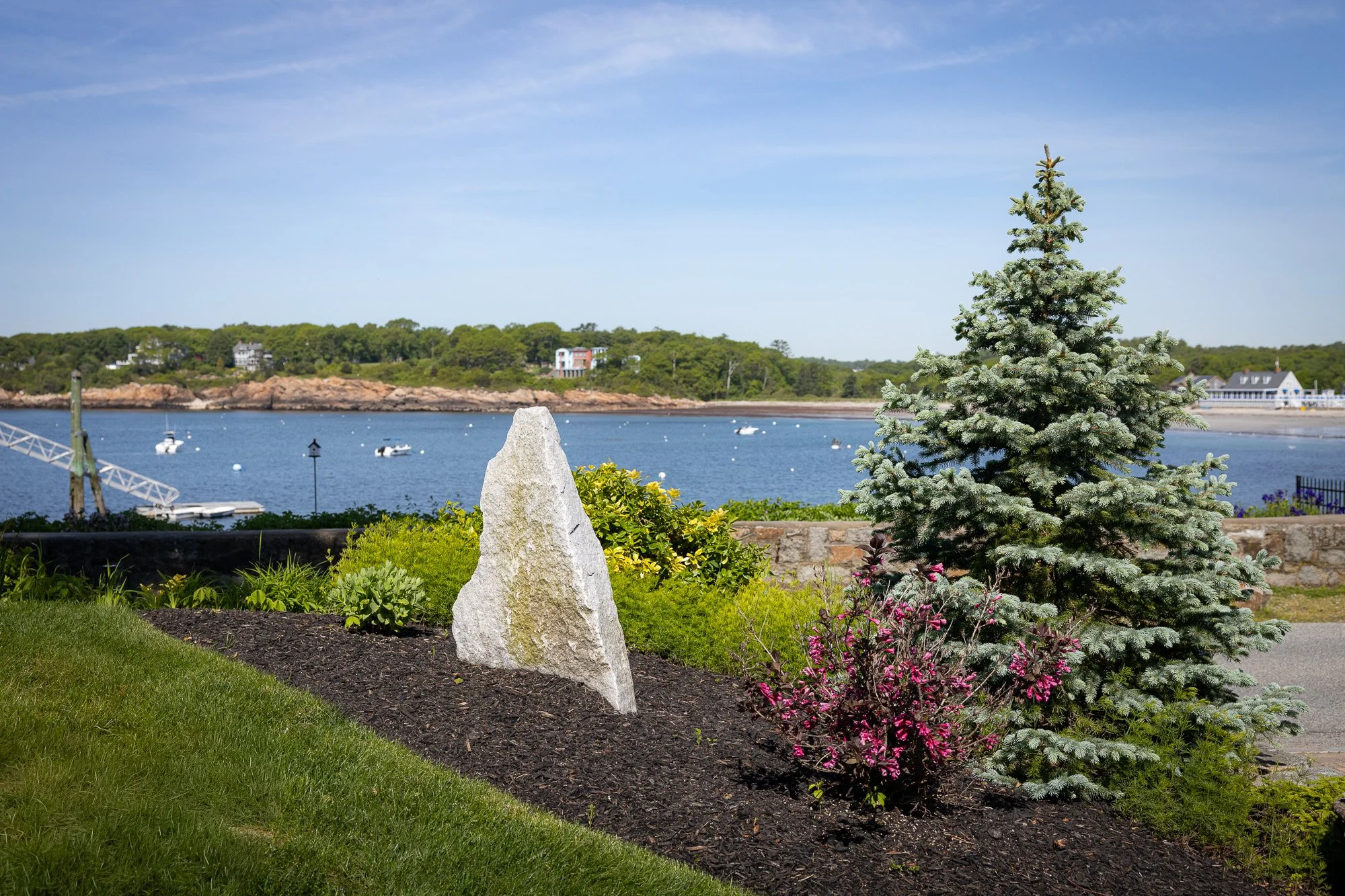 A landscaped yard by a body of water with boats, a small dock, and houses in the background. There is a blue spruce tree, pink flowers, a stone ornament, and a green lawn.