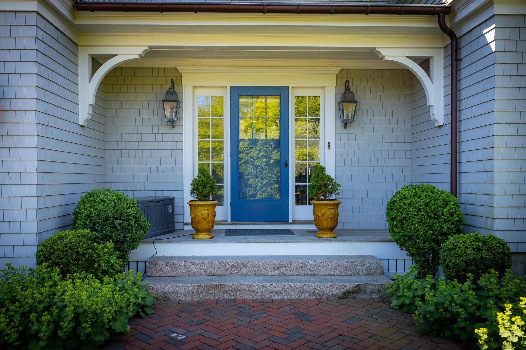 Front porch of a house with a blue door, two large potted plants, surrounded by green shrubs, and a brick pathway.