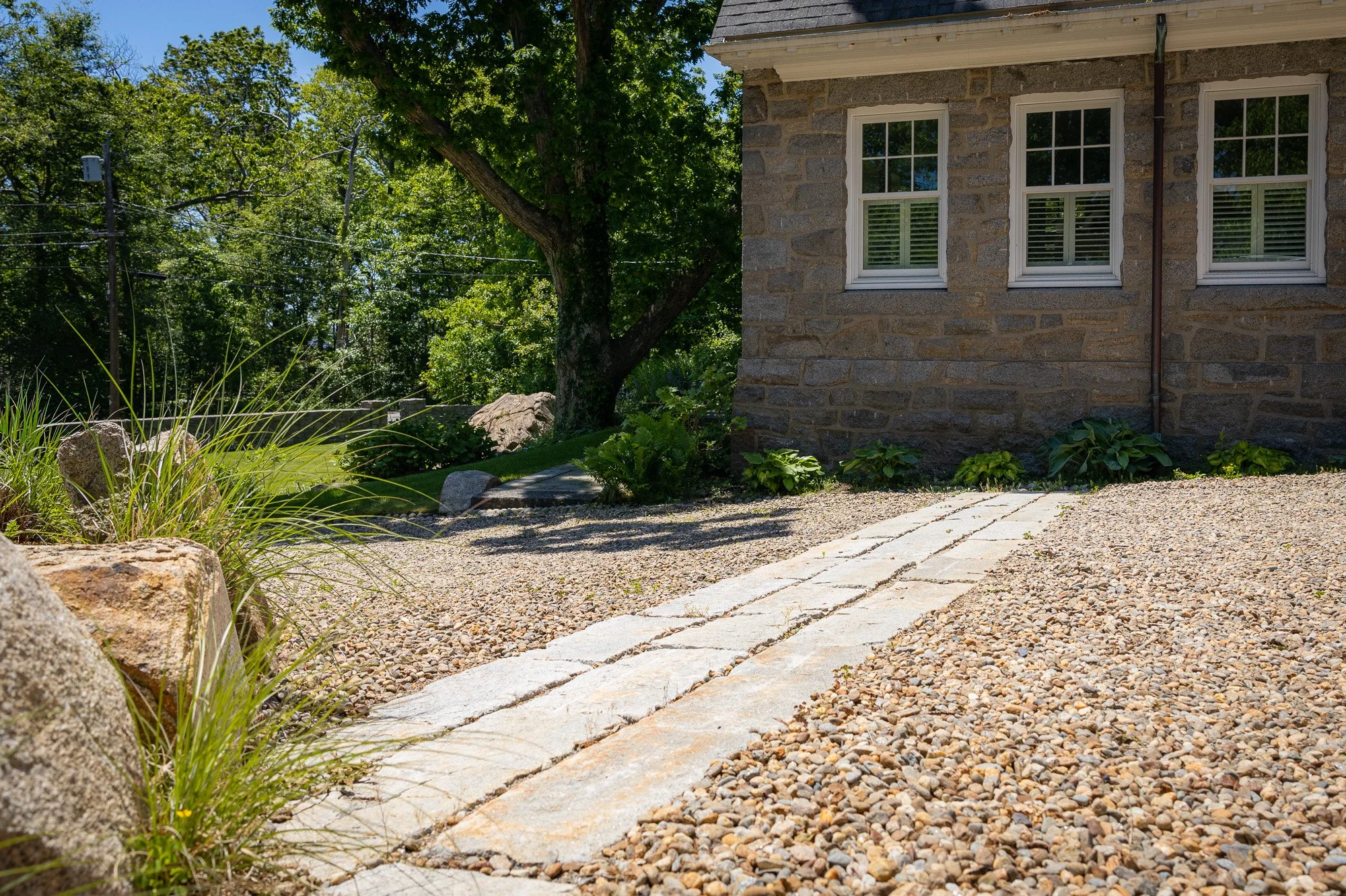 A stone walkway leading to the corner of a house with three windows, surrounded by gravel, plants, and trees, on a sunny day.