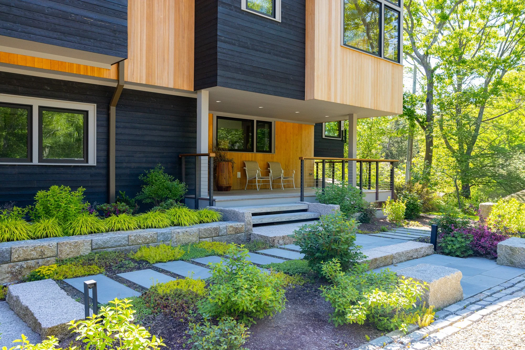 Modern house with a wooden exterior, large windows, and a small patio with chairs. The house is surrounded by lush greenery, including trees, bushes, and a well-maintained flower bed with yellow and purple flowers. Stone steps lead up to the patio.