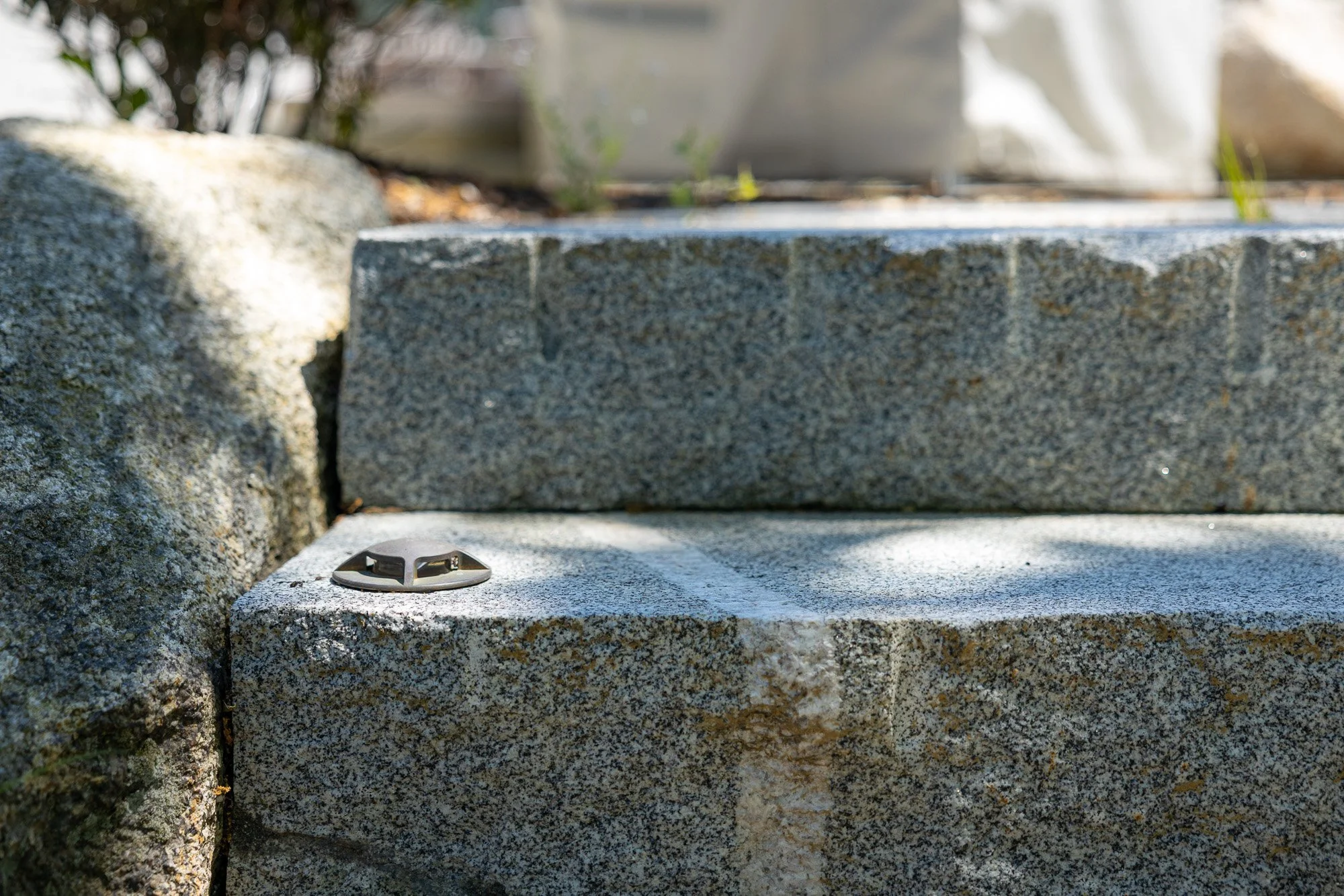 Close-up view of granite stairs with a metal stair tread on the bottom step, surrounded by stone and some greenery in the background.