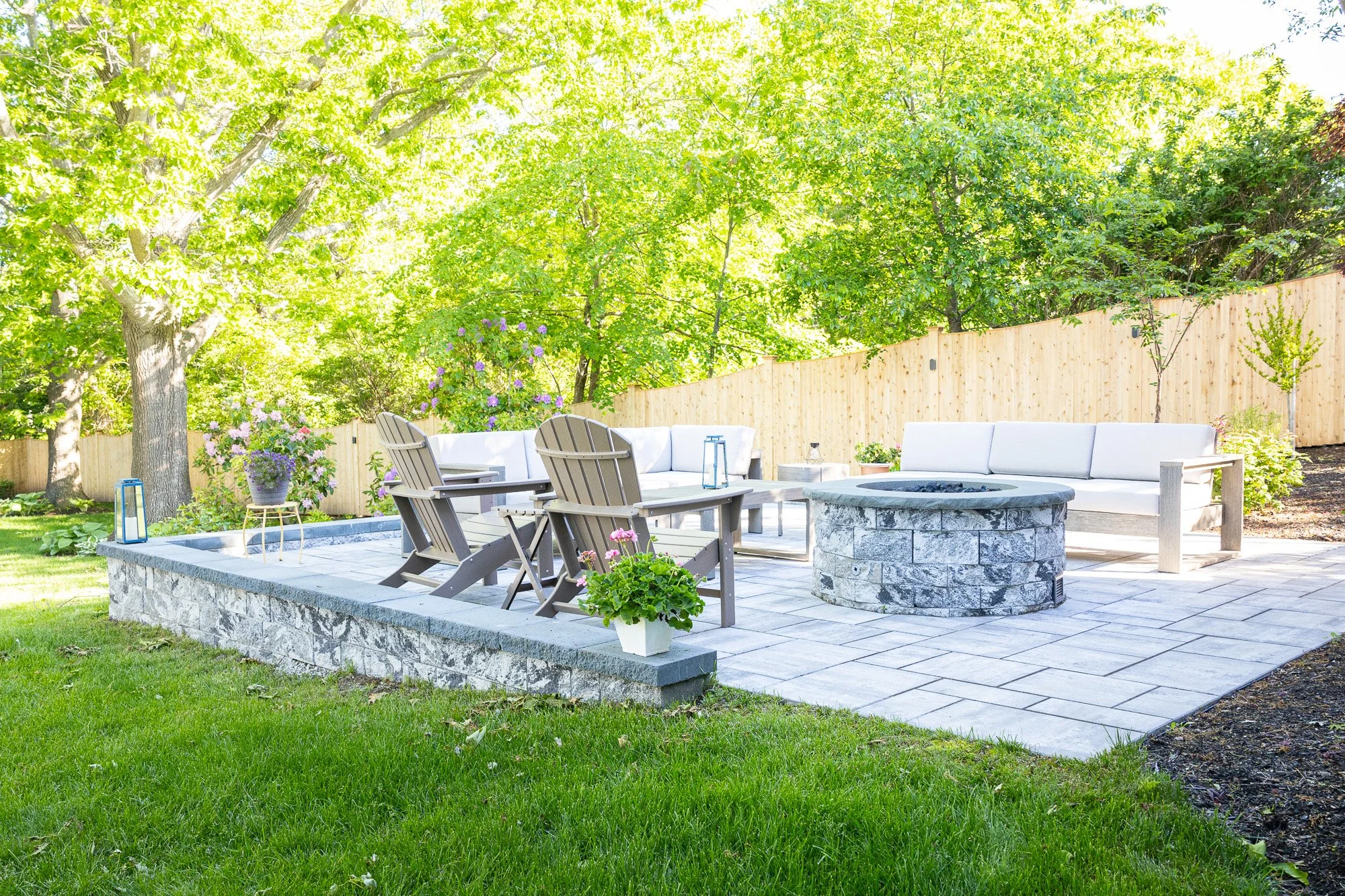 A backyard patio area with outdoor furniture, including white cushioned seating and wooden chairs around a fire pit. There are potted plants, lanterns, and a stone-edged platform with a striped flowerpot. Behind, there is a wooden fence and lush gree