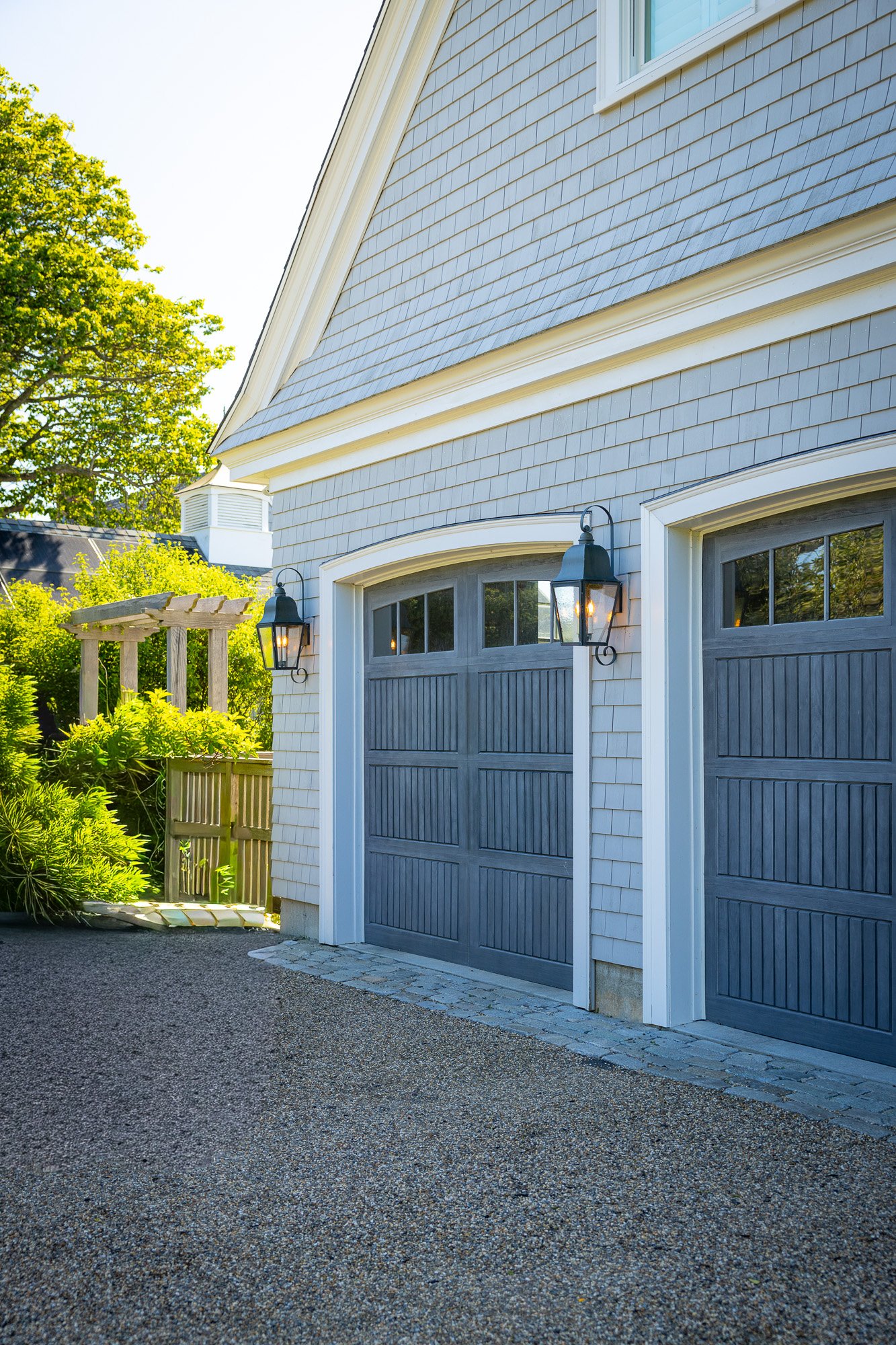 A house with gray garage doors and exterior sconces, surrounded by greenery, with a gravel driveway and a wooden arbor in the garden.