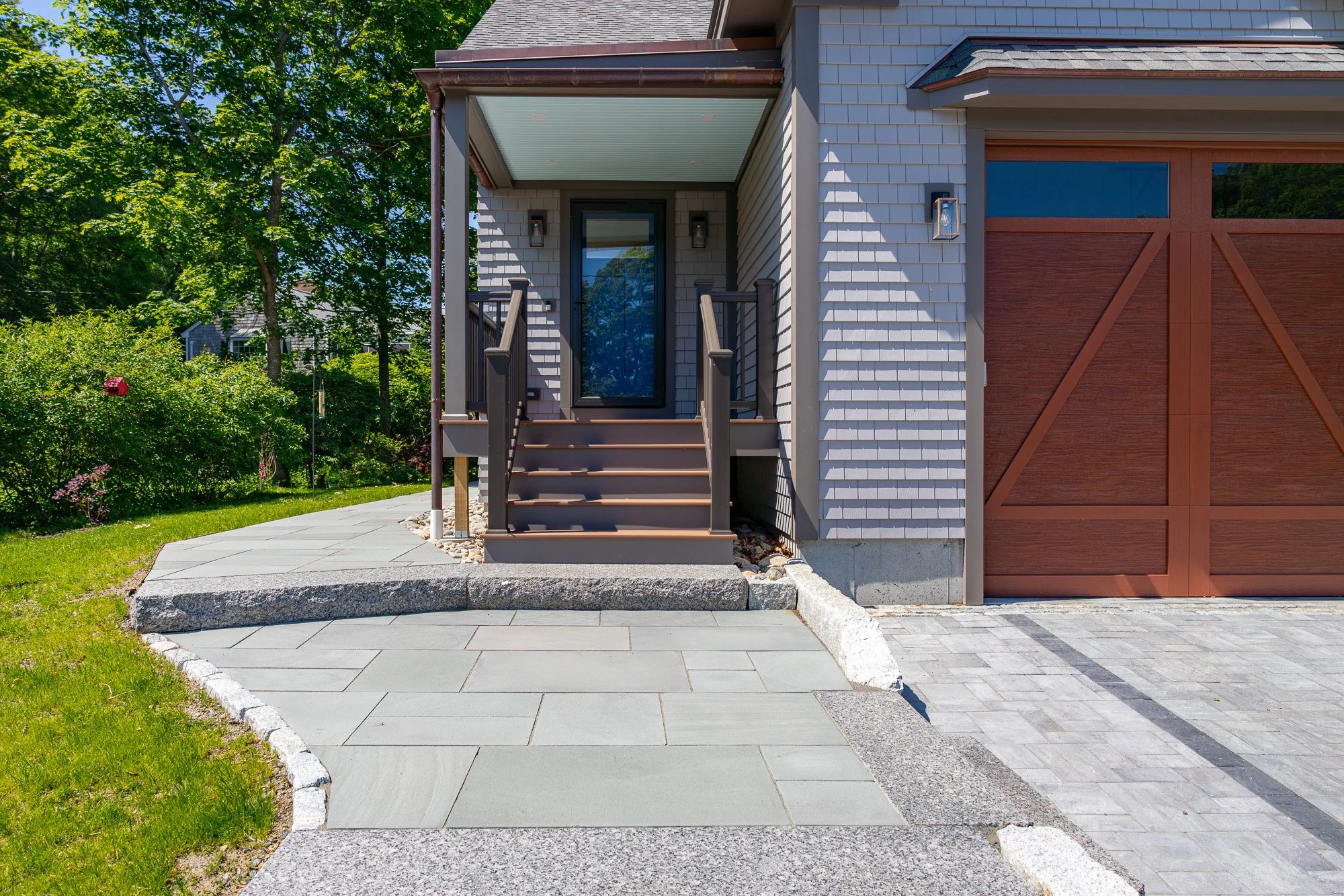 Front porch of a house with stairs leading up to a black door, a large window to the right, and a wooden garage door to the far right. The house has gray siding and a covered porch with a ceiling, surrounded by greenery, trees, and a garden.