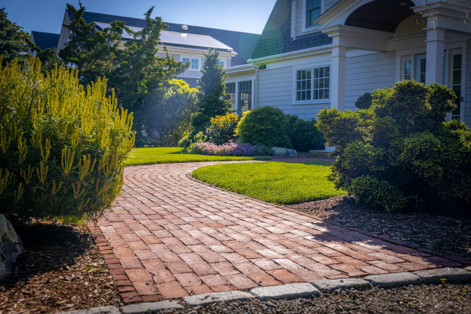 A brick pathway curving through a well-maintained lawn and garden with shrubs and flowering plants in front of a house with white siding and multiple windows, sunlight shining.