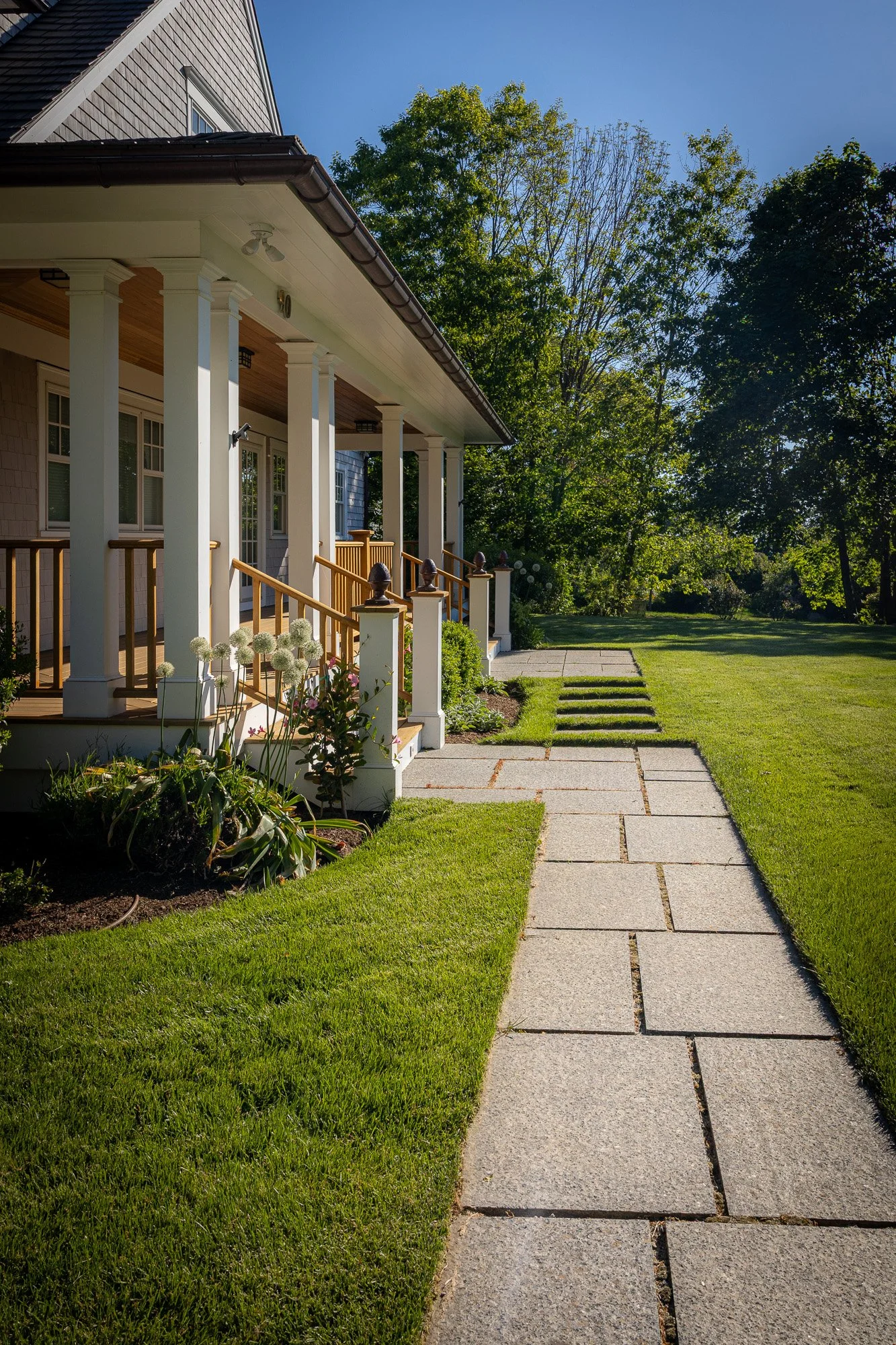 A front porch of a house with white columns and a wooden railing, a walkway with pavers leading to the sidewalk, and a green lawn with trees in the background under a clear blue sky.
