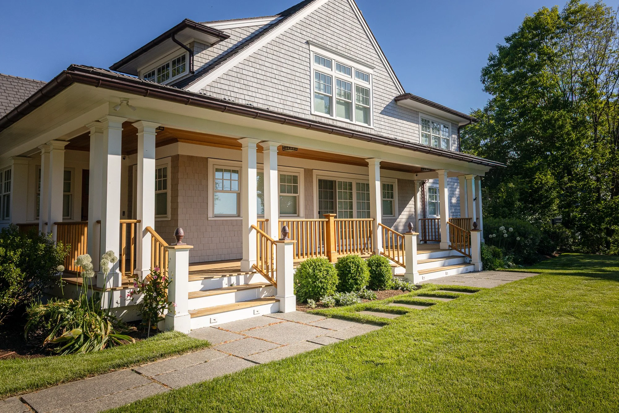 A large two-story house with a porch, wooden railings, and decorative columns. The house has a gray shingled exterior and multiple windows, some with white framing. There are landscaped plants and a sidewalk in front, with a blue sky and trees in the
