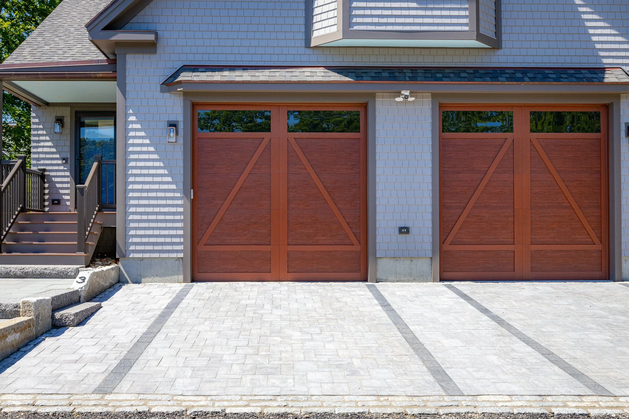Front view of a modern house with a driveway, two garage doors made of wood, gray brick and siding exterior, and small steps leading to a side door with an outdoor light fixture.