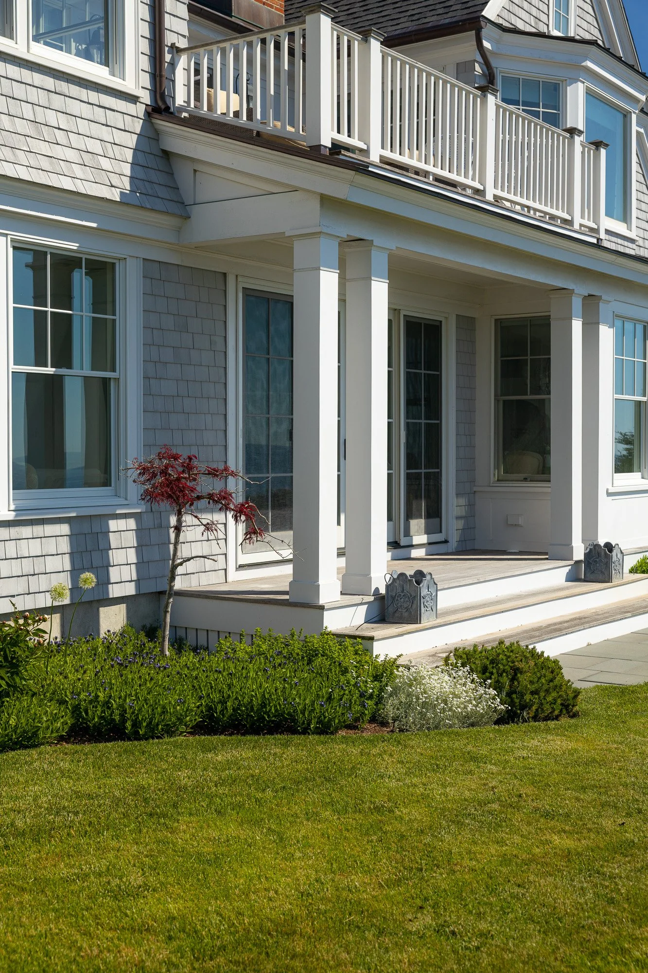 Front view of a multi-story house with a porch, white railing, and large windows, surrounded by a well-maintained garden with green grass and flowering plants.