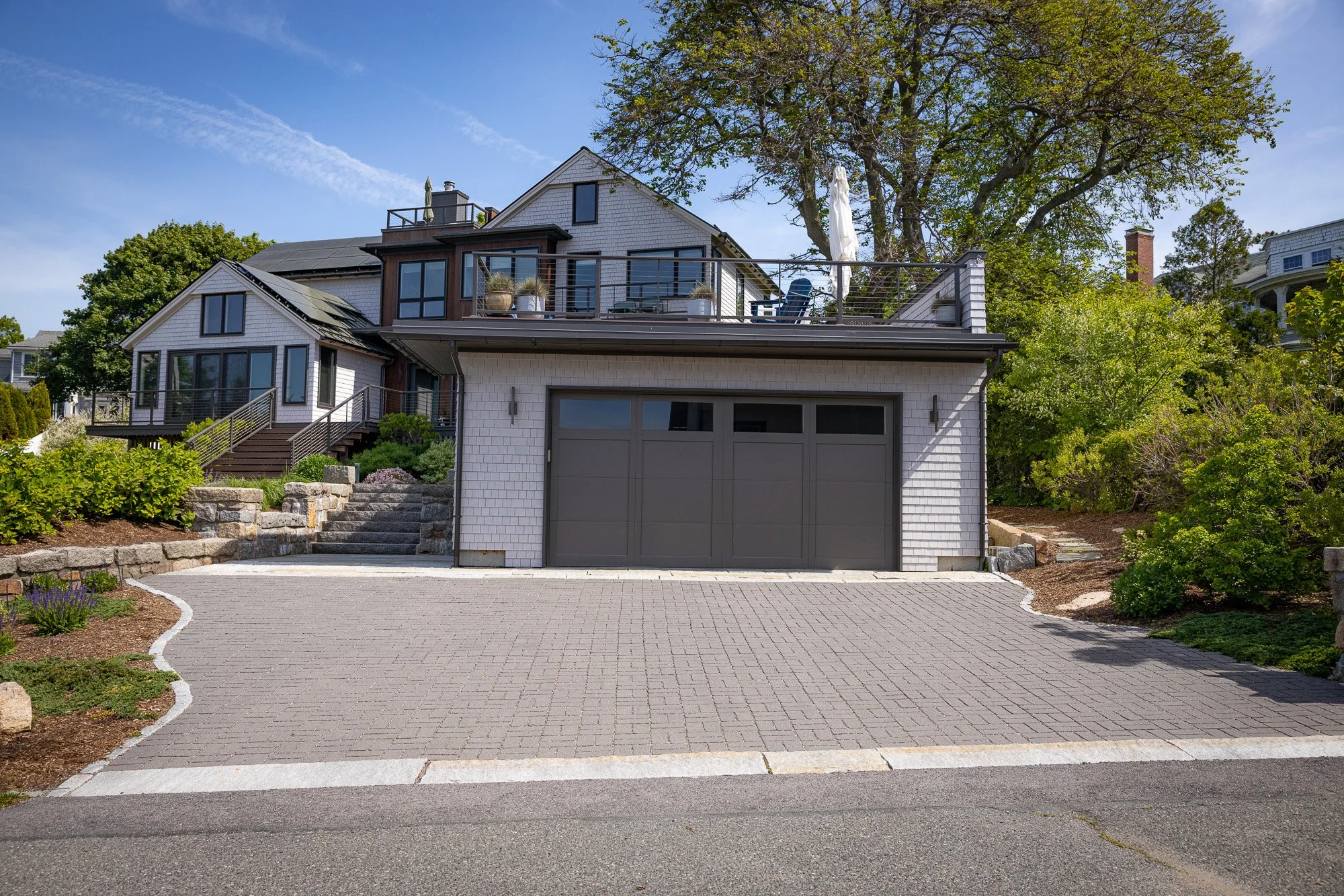 The image shows a modern house with a gray garage door and a drive parking area made of gray brick pavers. There are stairs leading up to a deck with outdoor seating, surrounded by plants and shrubs. The house has multiple levels, with large windows,
