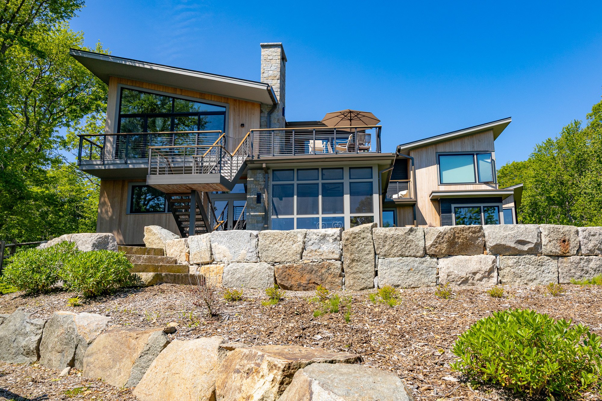 Modern house with large windows and a rooftop deck with outdoor furniture and an umbrella, surrounded by green trees and a stone retaining wall in the foreground.